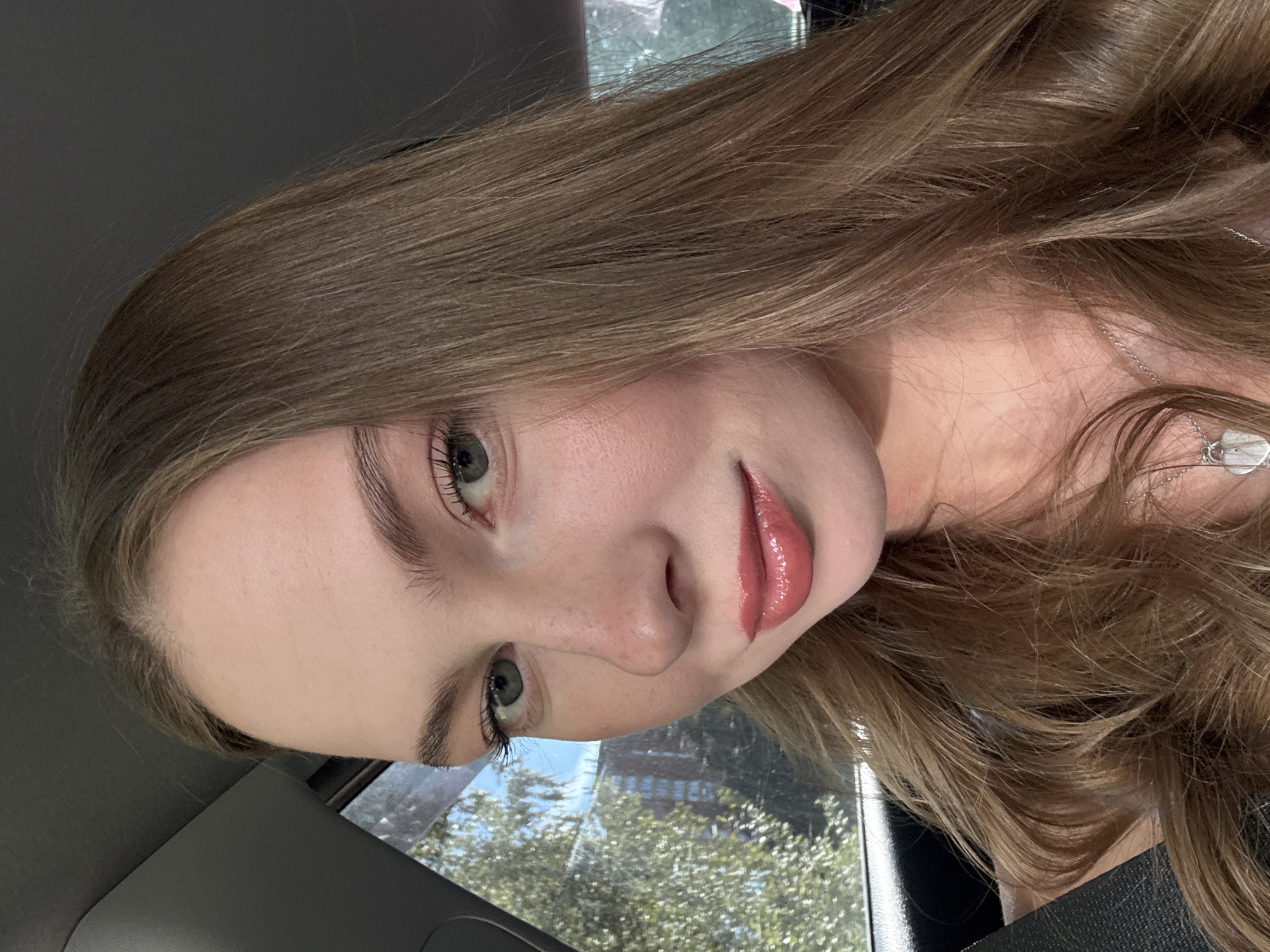 A young woman with light brown hair and green eyes is seen in a close-up shot, likely taken inside a vehicle. She is wearing glossy pink lipstick and has a subtle blush on her cheeks. Her hair is long and wavy, framing her face. The background is blurred, but suggests an outdoor environment with greenery and buildings visible through a window, hinting at a possible urban setting. The lighting appears to be natural daylight.