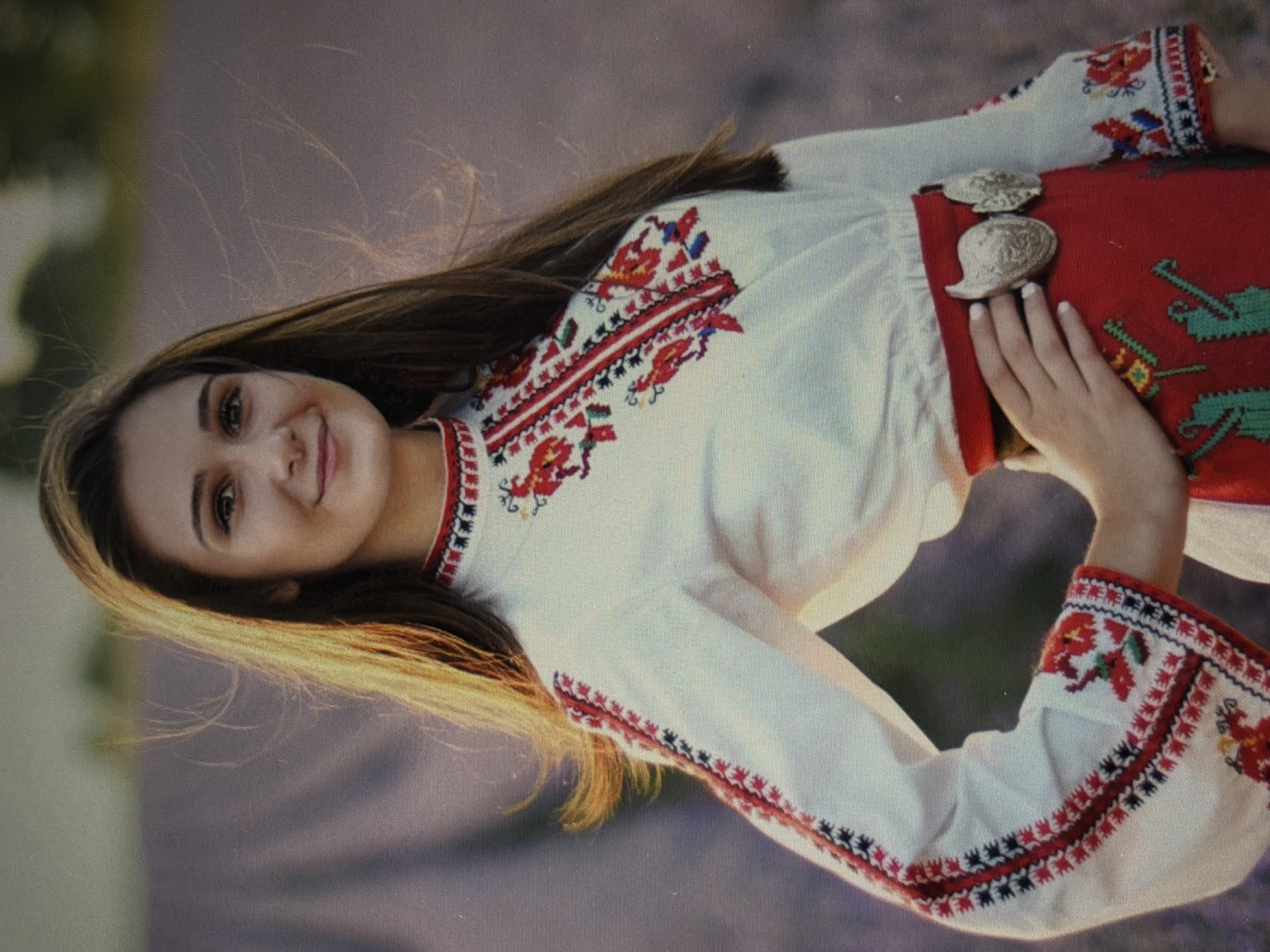 A young woman, with long brown hair and brown eyes, is pictured outdoors. She is wearing a white embroidered blouse with red and black patterns on the sleeves and neckline, and a red embroidered skirt with a silver belt. The woman is standing in what appears to be a field of lavender, with a blurred background of greenery and purple flowers. Her left hand is placed on her hip, and she is looking at the camera with a slight smile. The lighting suggests it is either dawn or dusk, with the sun casting a warm glow on her hair and face.