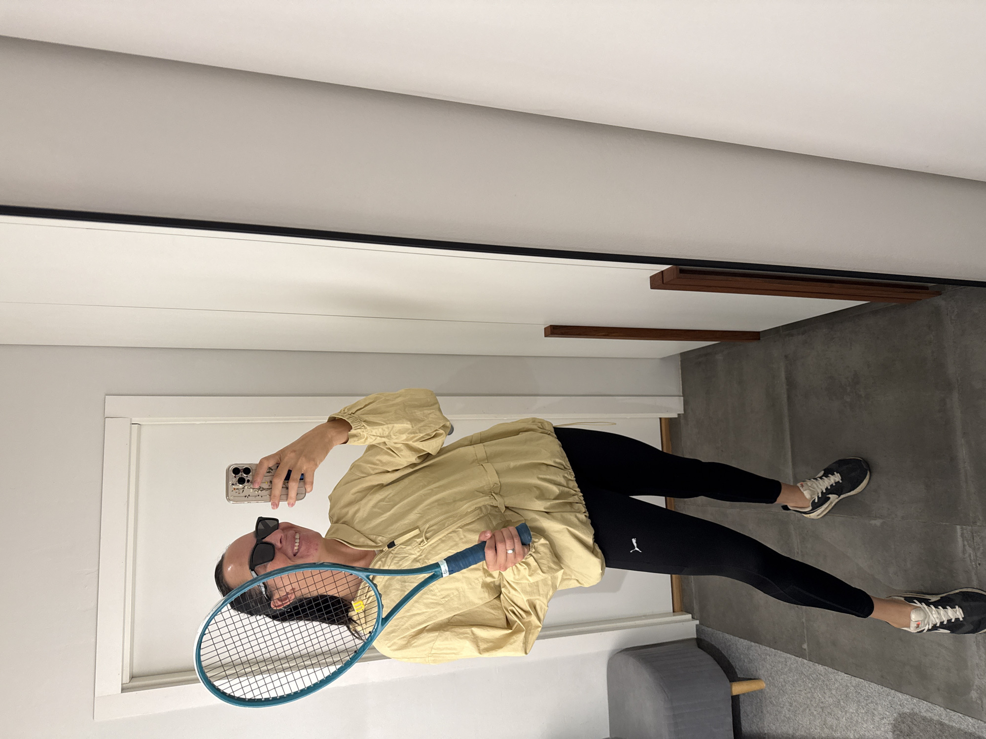 A woman is taking a selfie in what appears to be a dressing room or changing area. She is holding a tennis racket and wearing sunglasses, a light beige jacket, black leggings with a Puma logo, and sneakers. The background includes a door, a full-length mirror, and part of a gray upholstered stool. The floor is tiled with dark gray tiles. The woman's facial expression suggests a playful or confident mood. The image does not contain any elements that indicate the location is Chaikovsky, Russia.