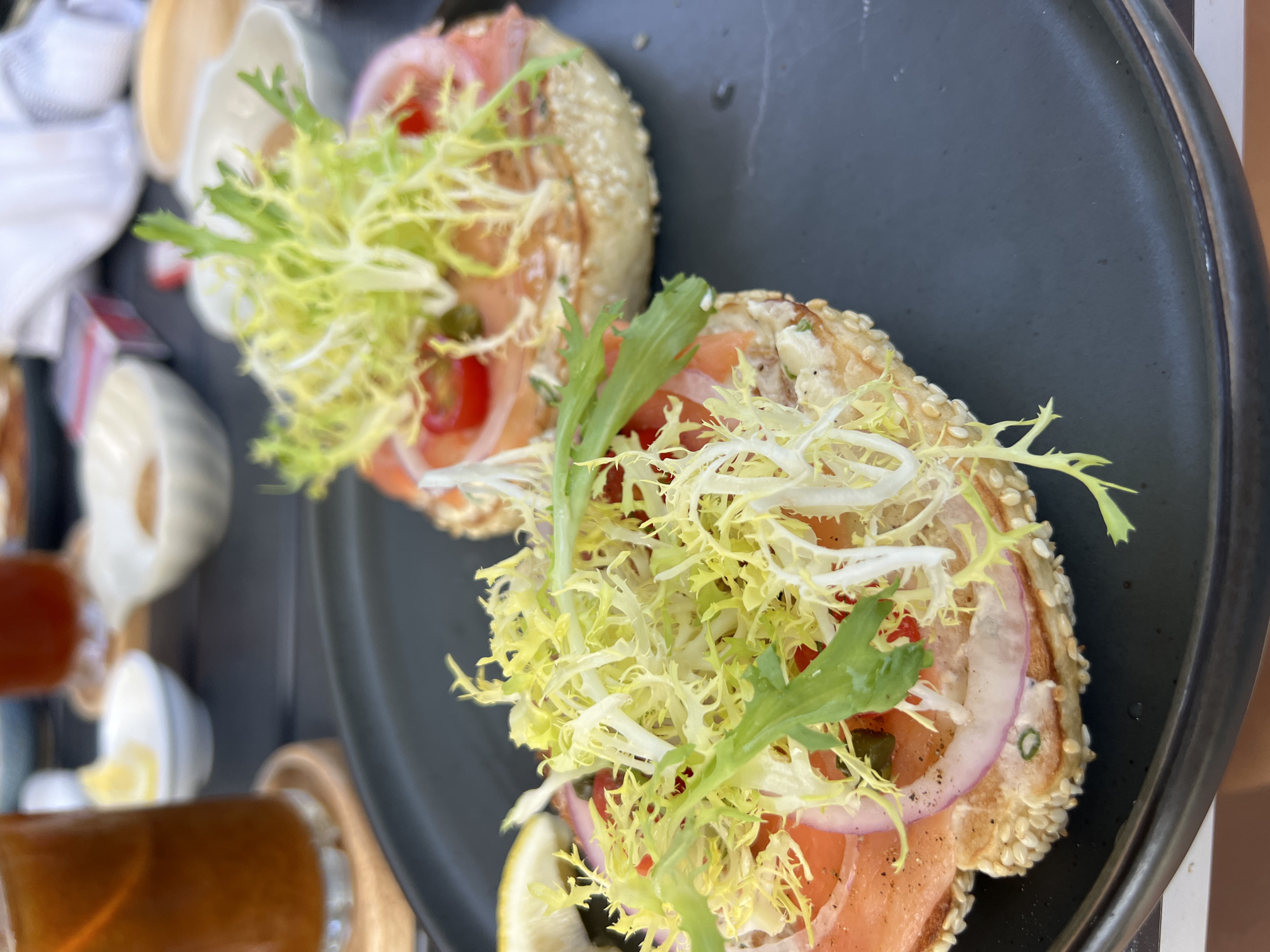 The image shows two open-faced bagels with smoked salmon, cream cheese, red onion, cherry tomato, and frisee lettuce on a black plate. A glass of iced tea and other dishes are visible in the background. The scene appears to be an outdoor dining setting, possibly at a cafe or restaurant in Choeng Thale, Thailand.