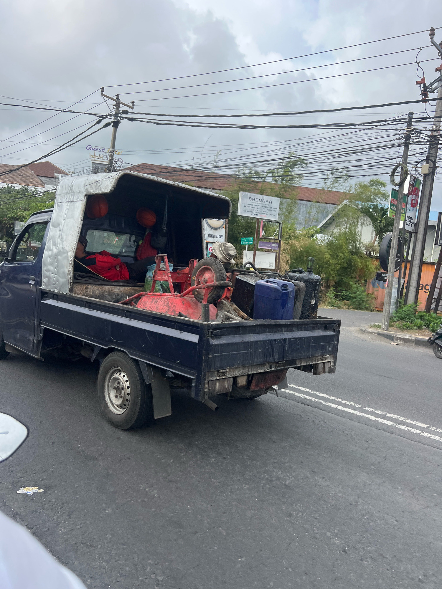 A dark blue utility truck is seen from the side and rear, traveling on an asphalt road in Denpasar, Indonesia, under a cloudy sky. The truck bed is filled with various items, including what appears to be a red lawnmower, several plastic containers (one blue, two black), and some red and blue fabric. Two orange spherical objects, possibly helmets or balls, are visible in the cabin. A person wearing red clothing is partially visible in the cabin, lying down. Utility poles with numerous wires are prominent in the background, along with buildings and lush green vegetation. Signs for "SASMARA" and "WISATA ATU BALI CAFE 2" are visible on the right side of the road. The truck's rear bumper shows signs of rust and damage. The road has double white lines marking the lanes.