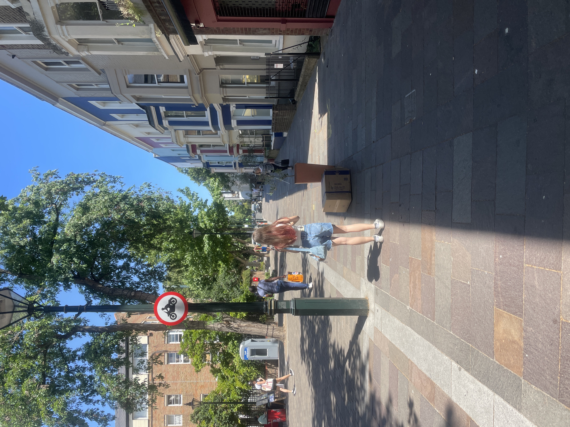 The image depicts a street scene in Notting Hill, London, United Kingdom, on a sunny day. A young woman with reddish-brown hair is walking away from the camera, wearing denim shorts, a white top, and sneakers, carrying a light blue bag. She is walking on a paved sidewalk made of multi-colored stone tiles. In the background, a row of colorful terraced houses line the right side of the street. The houses are painted in shades of blue, white, and grey, with large bay windows. Trees with green foliage line the left side of the street, casting shadows on the sidewalk. A traffic sign with a motorcycle symbol crossed out is visible on a lamppost. Other individuals are seen walking in the distance, and a blue ATM machine is present on the left. Cardboard boxes are placed on the sidewalk. The overall impression is of a vibrant urban environment on a clear day.