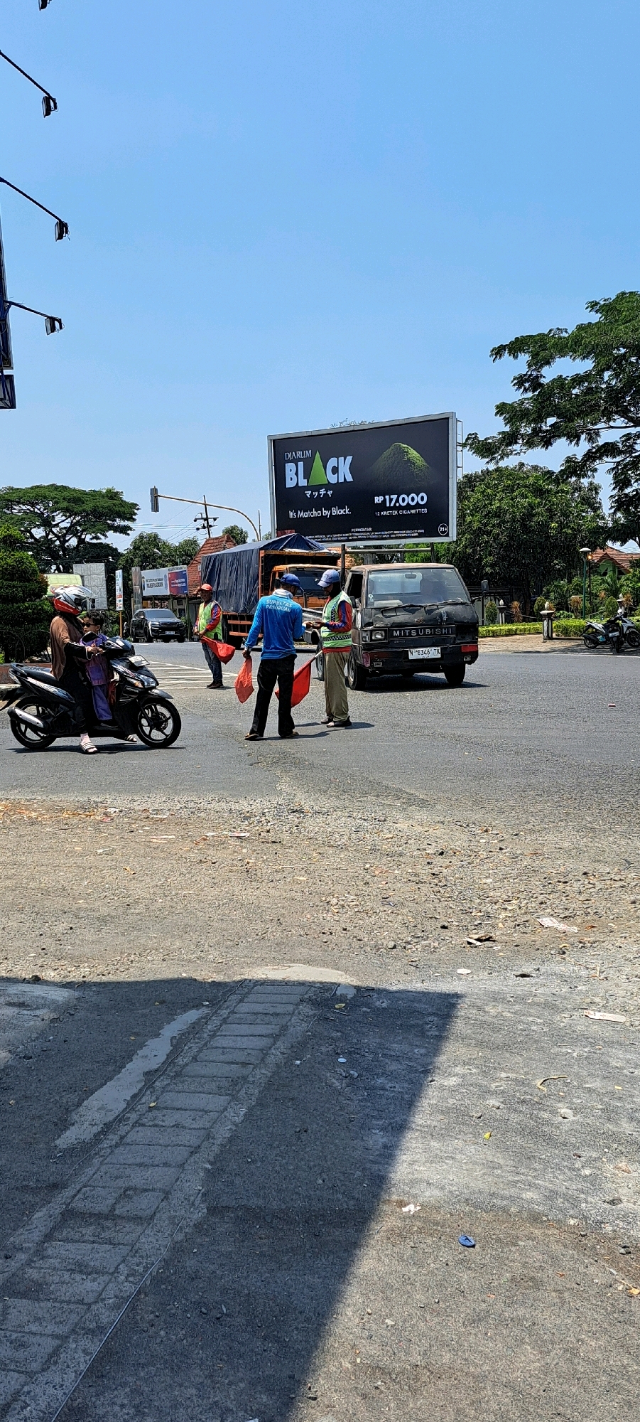 In Pandaan, Indonesia, under a clear blue sky, a busy intersection is depicted. A woman wearing a helmet and a purple top rides a scooter with a child behind her. Three individuals, likely traffic directors or workers, stand on the asphalt, each holding a red flag. One wears a blue shirt with "SUPERGAS PLUS GASPOLIM" printed on it, while the other two wear high-visibility vests over their shirts. Behind them, a large truck and a black Mitsubishi van are stopped. A billboard advertising "DIARUM BLACK MATCHA" is prominently displayed overhead. The ground is paved with asphalt and gravel, with a small drainage channel visible. The scene suggests a warm, sunny day with typical urban activity.