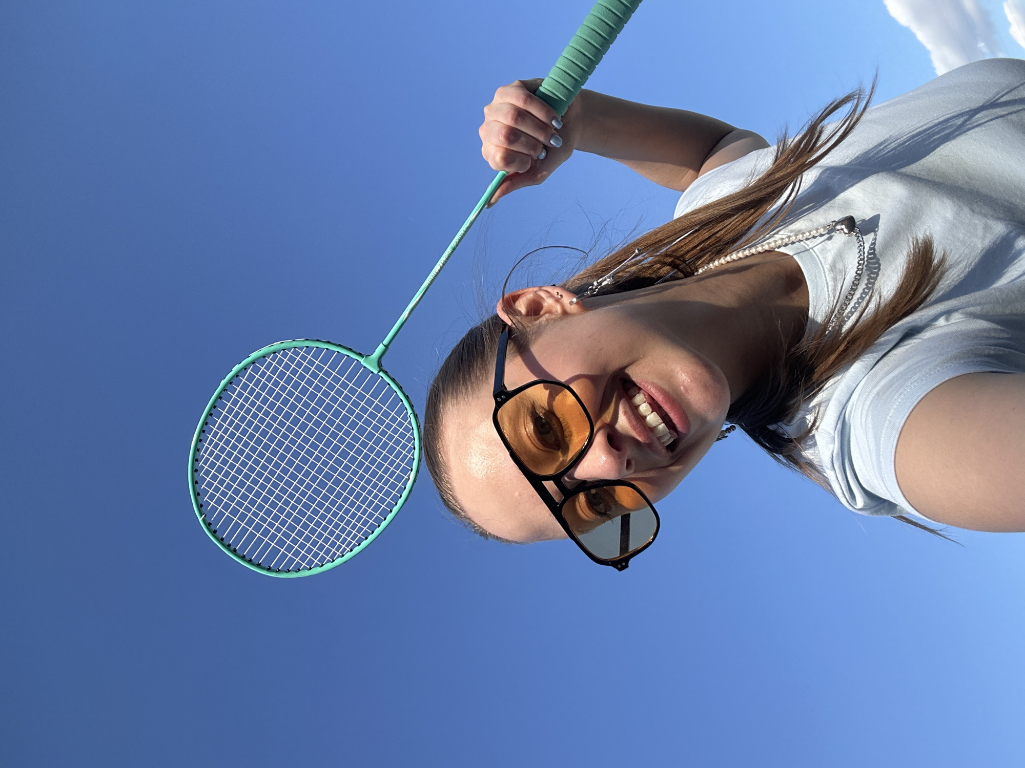 A young woman is captured in a selfie outdoors, holding a turquoise badminton racket. She is wearing a light blue t-shirt and sunglasses with orange lenses. A pearl necklace is visible around her neck. Her hair, long and brown, is styled down. She is smiling broadly, with her teeth showing, and her face is turned towards the camera. The background is a clear blue sky, suggesting a bright, sunny day. The image appears to be taken from a low angle, looking up at the woman. The orientation of the photo is such that the woman's head is at the bottom and her feet would be at the top, if they were visible. The scene is outdoors, with the sky dominating the frame, indicating it is likely daytime. There are some hints of clouds in the upper right portion of the image. There is no visible text in the image. The overall mood appears cheerful and playful, possibly indicating a recreational activity or a moment of enjoyment on a pleasant day in Khimki, Russia.