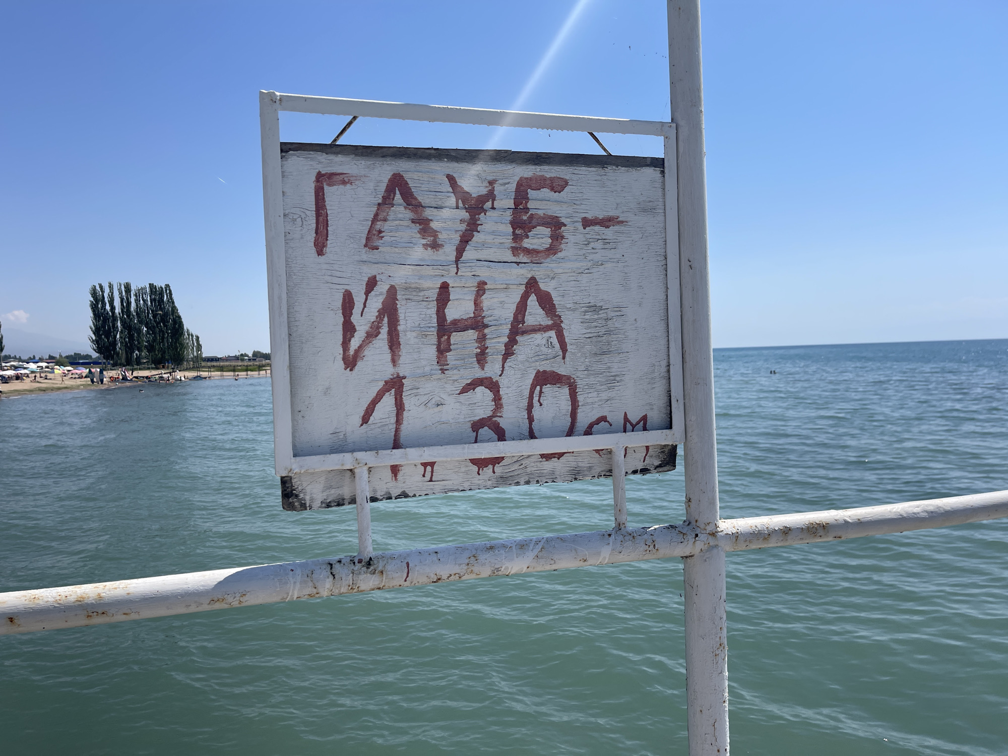 A weathered, white wooden sign with red lettering is mounted on a white metal post overlooking a body of water. The sign reads "ГАУБ- ИНА 1.30м". In the background, a beach is visible with numerous people and umbrellas. Tall, thin trees line the shore, and the sky is a clear blue. The water is a tranquil turquoise. The scene appears to be a public area, likely a beach or pier in the city of Ornek, Kyrgyzstan.