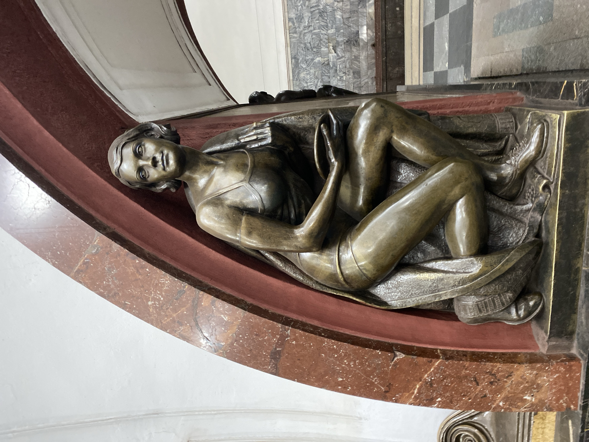 The image shows a bronze statue of a reclining woman, positioned on a reddish-brown marble base. The statue is located in the Moscow Metro, specifically at the station Ploschad Revolyutsii in Moscow, Russia. The woman is depicted wearing what appears to be sportswear and shoes and she is holding an object. Her posture is relaxed as if lounging. The backdrop includes a wall with gray tones. The statue is surrounded by architectural features.