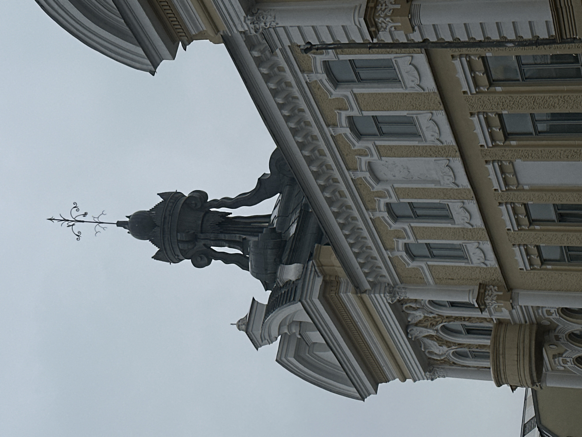 The image shows a low-angle view of the roofline of a building, probably located in Kazan, Russia, as indicated by the architecture. The building features a sculpture of a winged figure with a spear-like ornament at the top, set against a gray sky. The building itself is detailed with ornate window frames, decorative moldings, and columns. The color palette includes beige, cream, and gray tones, typical of historical architecture in that region. The photo appears to have been taken on an overcast day.