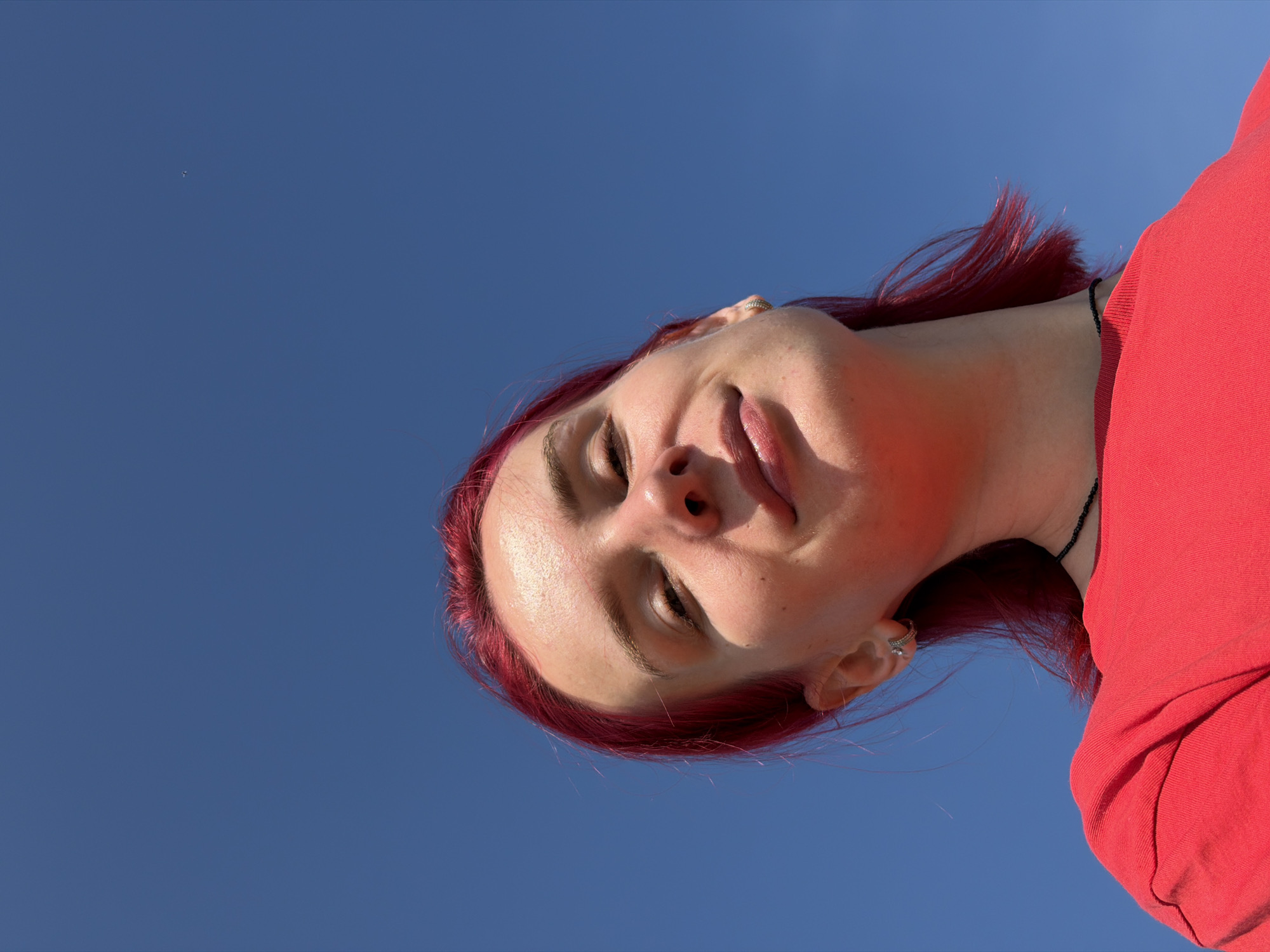 A young woman with bright red hair and fair skin is captured from a low angle against a clear blue sky. She is wearing a vibrant red t-shirt and a thin black beaded necklace. Her head is tilted back, and she is looking upwards with a slight smile. The sunlight casts a warm glow on her face, highlighting her features. An earring is visible in her left ear. The background is entirely composed of the vast, cloudless sky, suggesting an outdoor setting. There are no discernible city landmarks or any indication of the city of Chaikovsky, Russia, within the frame.