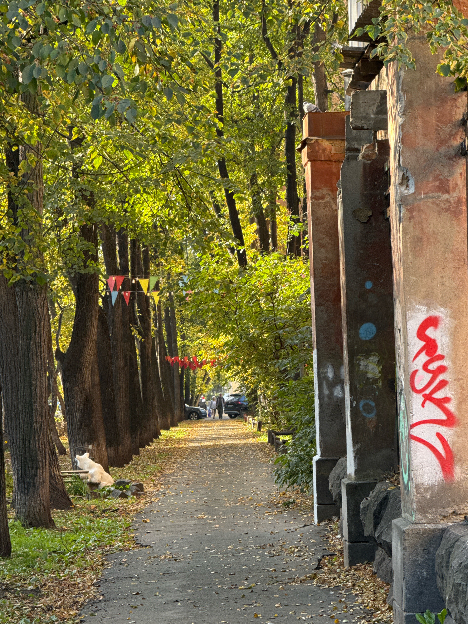 The image depicts a pathway lined with trees on the left and a dilapidated wall on the right. The pathway is paved and covered with fallen leaves, suggesting an autumn setting. Along the trees, a string of colorful triangular flags is hung. In the distance, several cars are parked and two individuals are walking. On the left side of the pathway, near the base of a tree, a light-colored dog is lying down. The wall on the right is weathered and covered in graffiti, with a prominent red tag. A pigeon is perched on a ledge of the wall in the upper part of the image. The scene appears to be an urban environment, possibly a park or a street with old architecture. This location is identified as an alleyway in the city of Yekaterinburg, Russia, near the historical building at 7 Vokzalnaya Street.