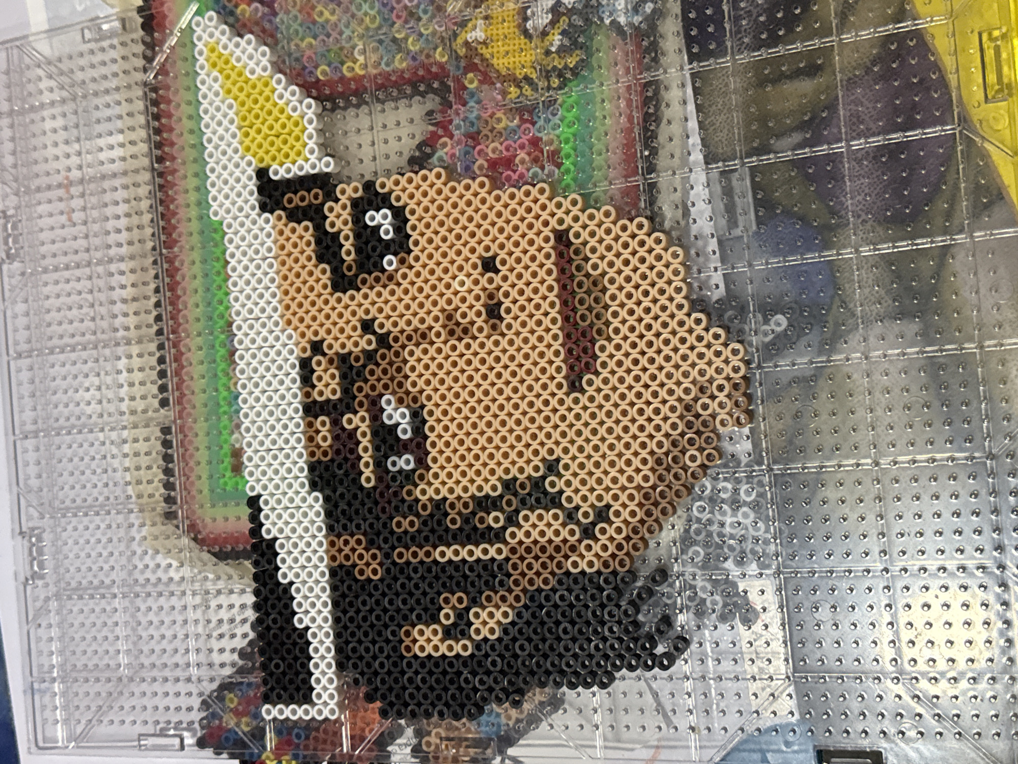 The image captures a close-up view of a perler bead artwork resting on a clear, grid-patterned pegboard, likely in Puebla, Mexico. The primary subject of the artwork is a stylized character, possibly a man, crafted from numerous small, cylindrical plastic beads. The character exhibits a light brown complexion, defined by light brown beads, along with black beads forming prominent hair or a beard, and facial features such as eyes. White and yellow beads create details around the top and side of the character's head, suggesting a hat, crown, or stylized element.
The perler bead creation is centered on the transparent pegboard, whose grid structure is clearly visible. Scattered around the edges of the artwork, particularly on the lower left and right, are various loose perler beads of different colors, including red, green, yellow, blue, black, and white, indicating that a crafting activity has recently occurred or is still in progress. The background beyond the pegboard is intentionally blurred, showing hints of other colors and abstract patterns, consistent with an indoor setting such as a crafting area or table. There is no visible text legible within the image.