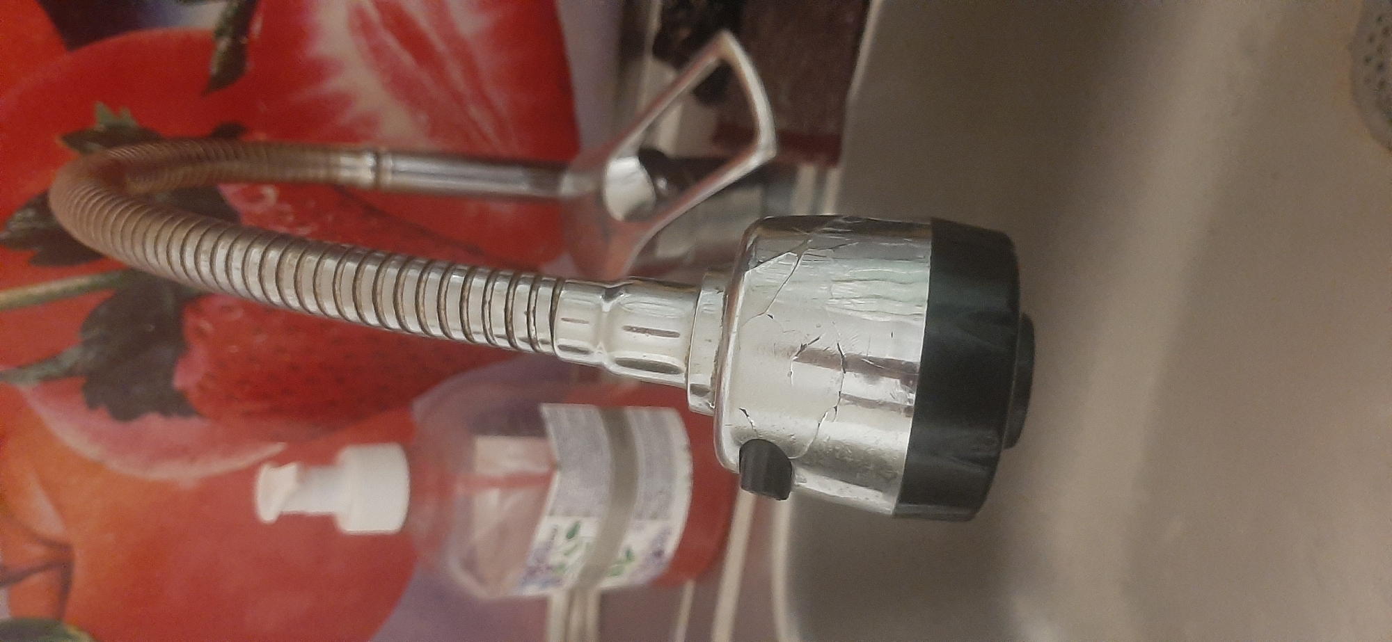 A close-up shot shows the head of a kitchen faucet with a flexible gooseneck. The faucet head is silver and has a black switch or button on the side. The faucet is positioned over a stainless steel sink. In the background, a clear plastic bottle of pink liquid soap with a white dispenser is visible, as is a wall covered with a pattern of red strawberries and green leaves. The scene is likely indoors, in a kitchen or bathroom. No specific location in Yessentukskaya, Russia, can be determined from the image.