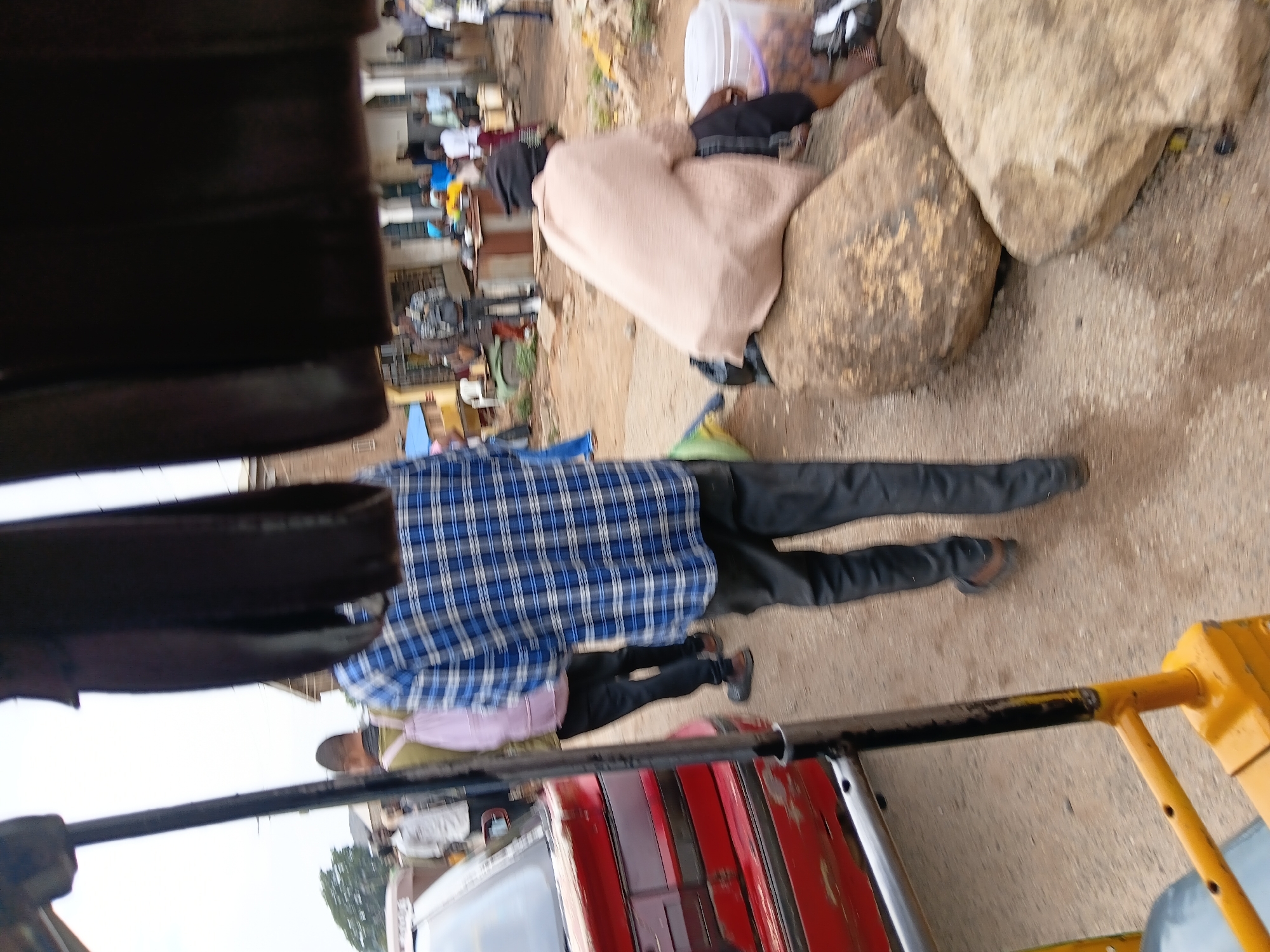 A street scene in Gwong, Nigeria, shows people gathered outdoors. One person sits on a collection of large rocks, covered with a light tan cloth. Another person wearing a blue plaid shirt and dark pants walks on the ground. A red and white vehicle, potentially a bus or shared transport, is visible near the lower edge of the image with yellow supports. Other individuals can be seen in the background. The ground appears to be dirt or gravel.