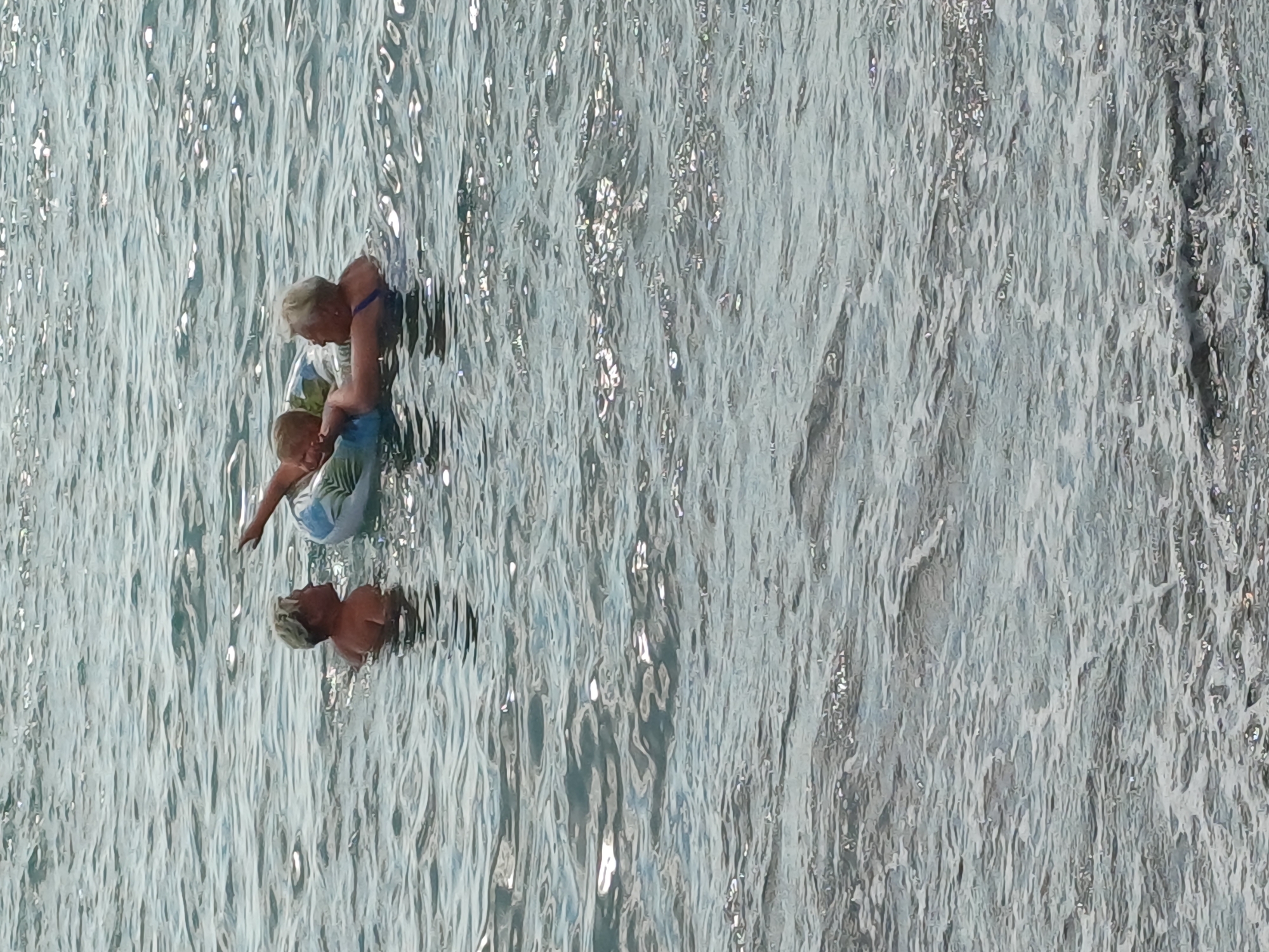 The media file shows a high-angle view of two people in shimmering, light-blue water, possibly a pool or the sea. The water is highly reflective, with bright highlights suggesting a sunny day. A woman with blonde hair is leaning forward, holding a child who is also blonde. The woman is wearing a blue strap on her shoulder and a patterned swimsuit or sarong. The child is facing away from the camera, with their head tilted back, and appears to be wearing a similar patterned garment. They are positioned in the upper left quadrant of the image. Below them and slightly to the right, a reflection of a person's head, also with blonde hair, is visible in the water. The overall impression is one of a calm, outdoor setting with recreational activity. The time of day appears to be daytime, and the weather is likely sunny, given the water's reflections. No text is visible in the media file. The context provided indicates the location is Sochi, Russia, which might imply a warm climate suitable for swimming or water activities.