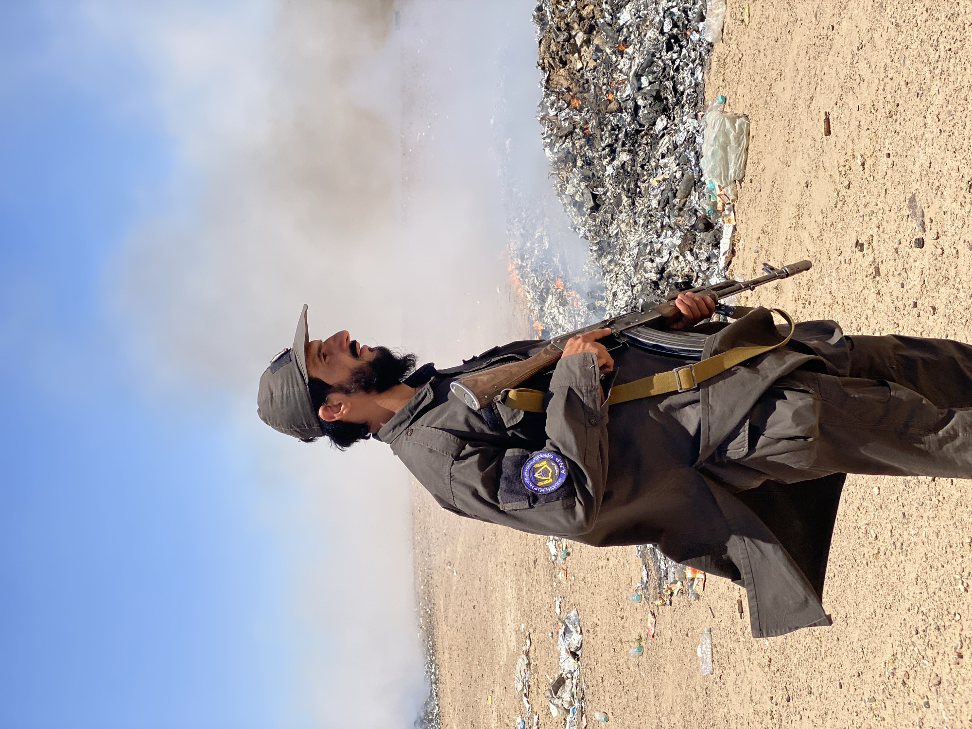 A man in a dark uniform and cap stands in a desert landscape, holding an assault rifle. Behind him, a large pile of burning trash emits smoke into the clear blue sky. The ground is littered with debris and plastic bottles. A patch on the man's left sleeve bears a circular emblem with Arabic script. The man has a beard and is looking to the right. The overall scene suggests a remote, possibly rural, location. The man's uniform and the presence of a weapon indicate a security or military role. The burning trash suggests a waste disposal site or an uncontrolled burning of waste. The environment is dry and dusty, typical of a desert or arid region.