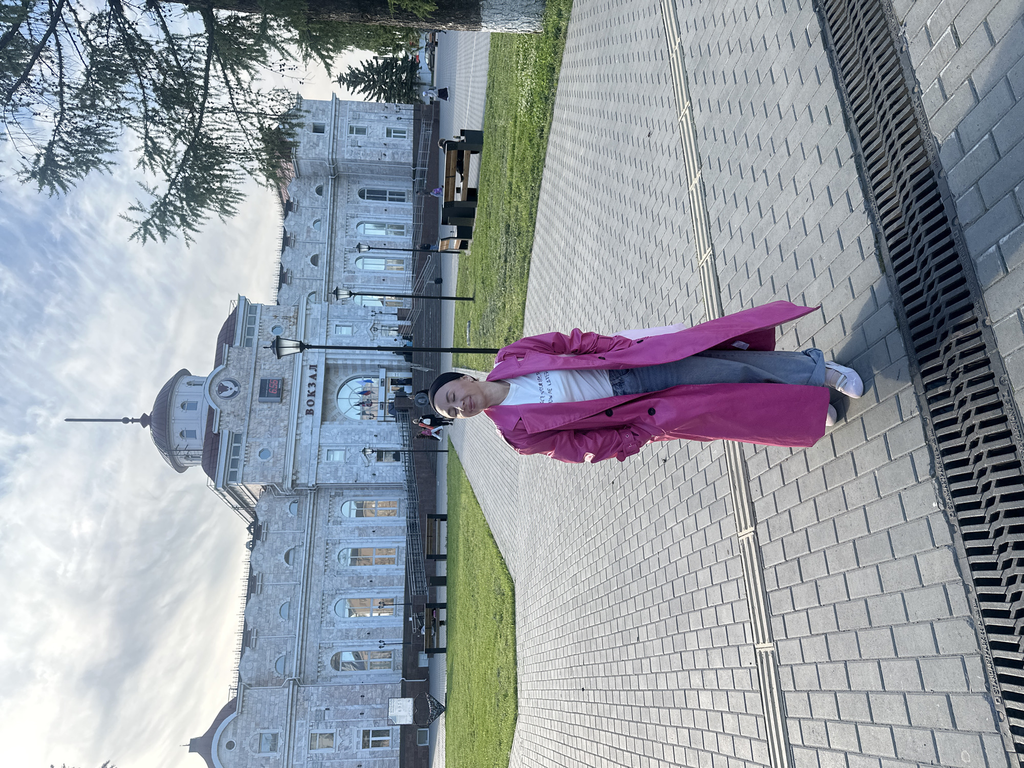 The image captures a person standing outdoors on a paved walkway, with a majestic train station building in the background. The scene is set in Izhevsk, Russia. The individual, dressed in a bright pink trench coat, a white t-shirt, blue jeans, and white sneakers, with a dark cap, is positioned slightly to the right of the center, looking towards the viewer with a subtle smile.
The multi-story building behind them is constructed from light-colored stone, featuring classical architectural elements such as numerous arched windows, decorative cornices, and a prominent dome topped with a spire. The word "ВОКЗАЛ" (VOKZAL), Russian for "train station," is clearly visible on the building's facade, along with a digital clock displaying "6:55".
The paved walkway, composed of grey rectangular pavers, runs diagonally across the lower right portion of the frame. Adjacent to it is a strip of vibrant green grass, and further back, dark green trees line the area. A metal drainage grate is visible along the right edge of the paved path, bearing faint text that includes "STANDARD" and "2019". The sky above is partly cloudy, indicating daytime conditions. The overall atmosphere appears calm and bright, depicting a casual moment in a public urban space.