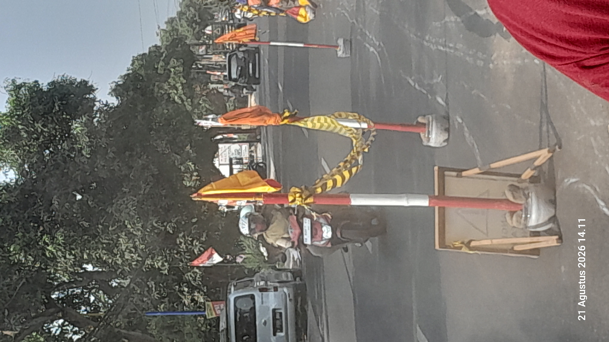 In the city of Takeran, Indonesia, on August 21, 2026, at 14:11, a street scene unfolds. A person wearing a helmet and a tan shirt rides a red motorcycle, navigating a road lined with trees. Red and white striped poles with yellow and orange flags are positioned on the side of the road, some adorned with black and yellow striped fabric. A silver van is parked on the left side of the image, while a dark-colored car drives in the background. In the foreground, a wooden easel-like sign is propped up, supported by what appear to be small rocks or weights. The overall environment suggests an outdoor urban setting with traffic and roadside markers.
