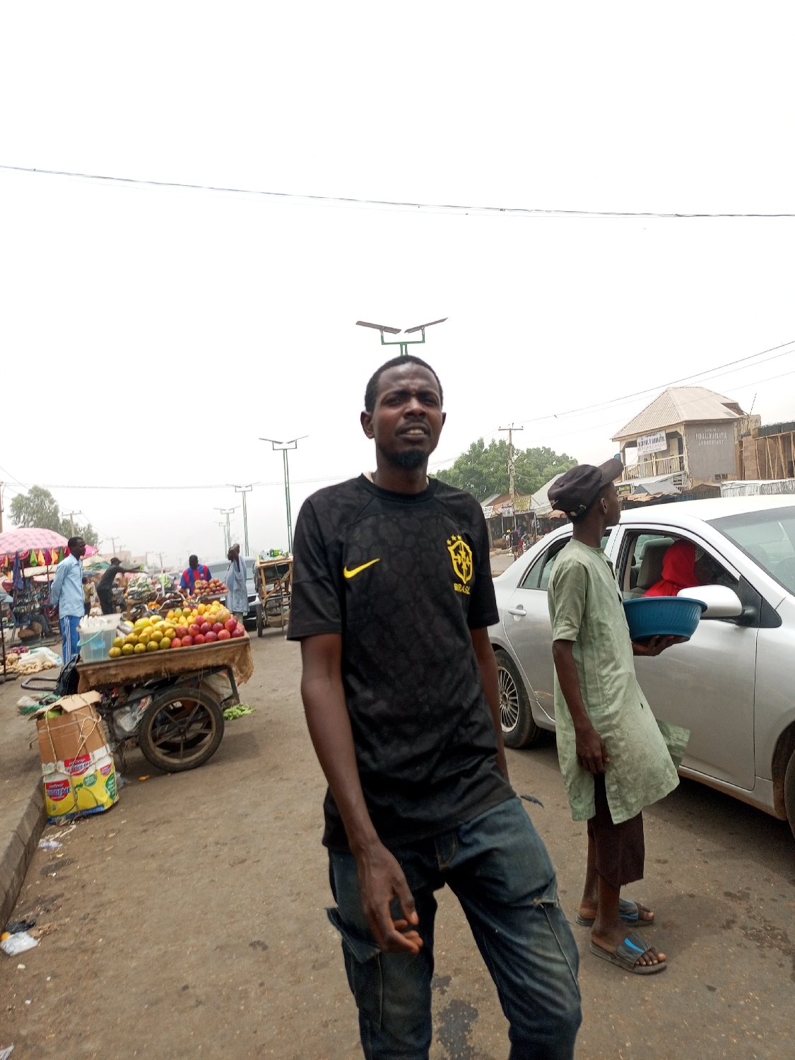 The image depicts a bustling street market scene in Tempure, Nigeria, during daylight hours under a bright, hazy, or overcast sky.
The primary subject is a dark-skinned man standing in the foreground, facing slightly to the right with a somewhat open mouth, as if speaking or observing. He is wearing a black t-shirt with a yellow Nike swoosh logo and the word "BRASIL" written in yellow on the chest. He also wears distressed blue jeans.
In the midground to the left, there is a wooden cart piled high with various fruits, predominantly yellow and red mangoes. Nearby, several cardboard boxes, including one with distinctive yellow packaging, are stacked on the ground. Further back, market stalls are visible with other individuals, some appearing to be vendors or shoppers.
On the right side of the frame, another individual, possibly a boy or young man, stands beside a silver sedan car. He is dressed in a light green tunic and dark pants, and holds a blue plastic basin. A person in red clothing is partially visible inside the car. In the background, multiple solar-powered streetlights are seen, along with a building that has "CAPITAL PROPERTIES" and "ADVERTISE" written on its facade.
The setting is an outdoor market area with an asphalt road, suggesting a common urban or semi-urban trading environment. The time of day is clearly daylight, but the high white balance in the sky suggests either very bright, diffuse light or a hazy atmosphere.
