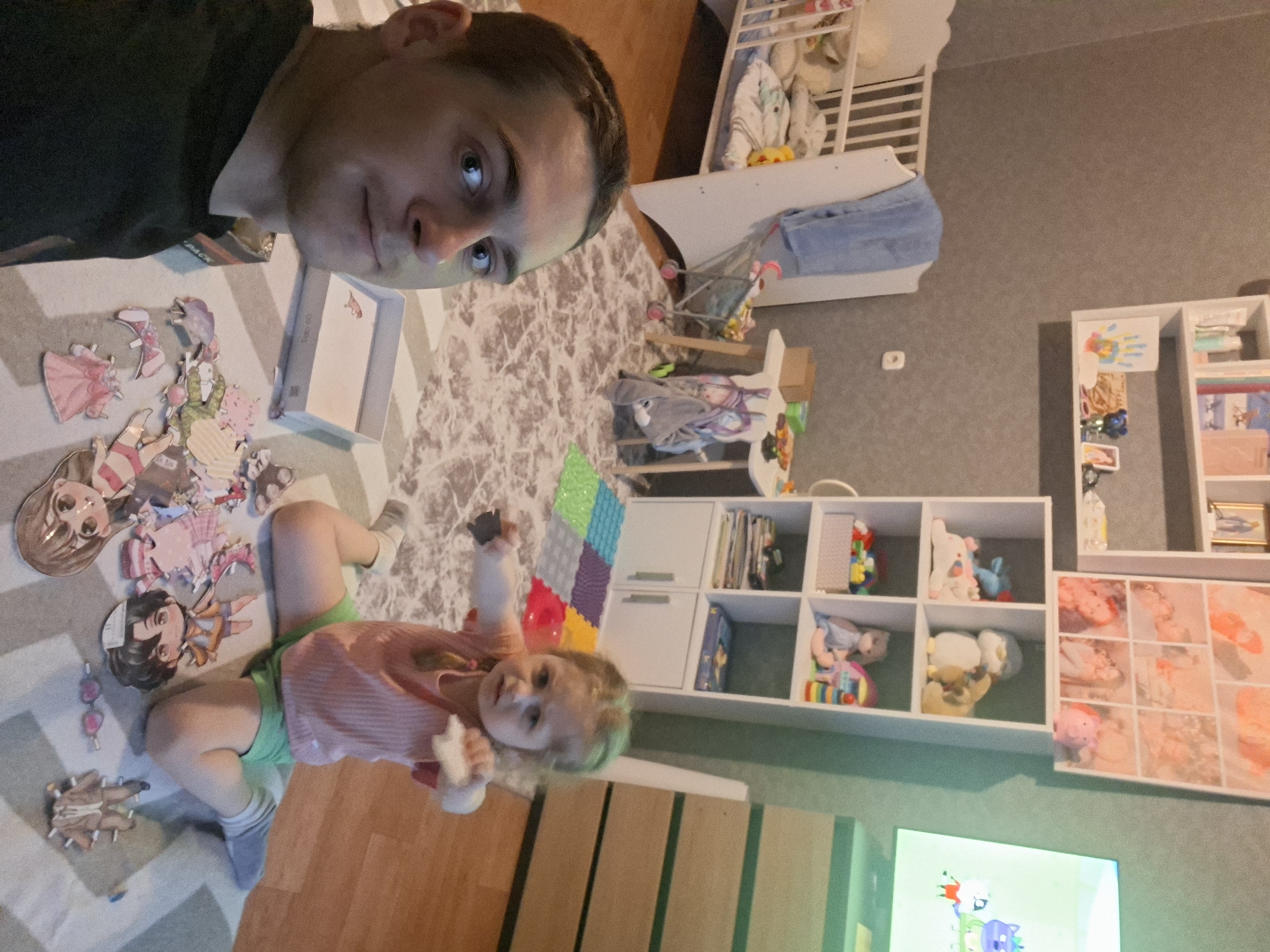 A man and a young child are on the floor in a room. The man, wearing a dark top, is looking at the camera. The child, wearing a pink and white striped top and green shorts, is holding objects in their hands. They are surrounded by paper dolls and accessories. The room contains a crib, white shelves filled with toys and books, and a TV playing a cartoon. The location is Unknown, Unknown.