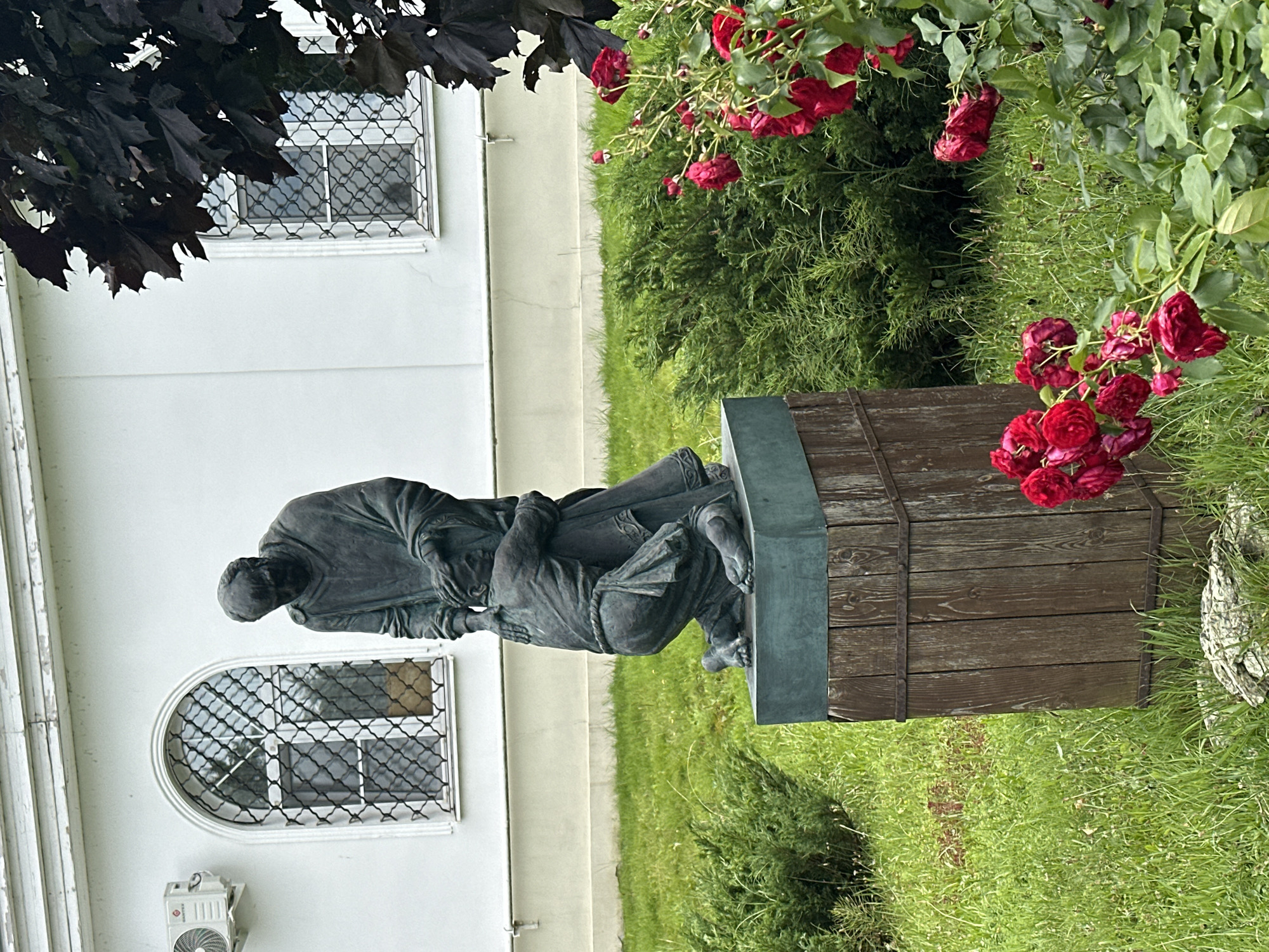 In Podolsk, Russia, a bronze sculpture depicts two figures in a tender embrace. The larger figure, standing and cloaked, gently places a hand on the head of the smaller figure, who kneels. The kneeling figure's face is bowed, conveying a sense of supplication or reverence. They are positioned on a wooden plinth with metal bands, set against a backdrop of a white building with arched windows. Lush green grass surrounds the sculpture, with a dense evergreen bush to the left and a blooming rose bush with vibrant red flowers in the foreground on the right. A dark-leaved tree extends from the upper right corner of the frame. An air conditioning unit is visible on the building to the left of the sculpture. The scene appears to be an outdoor public space, possibly a garden or park associated with the building.