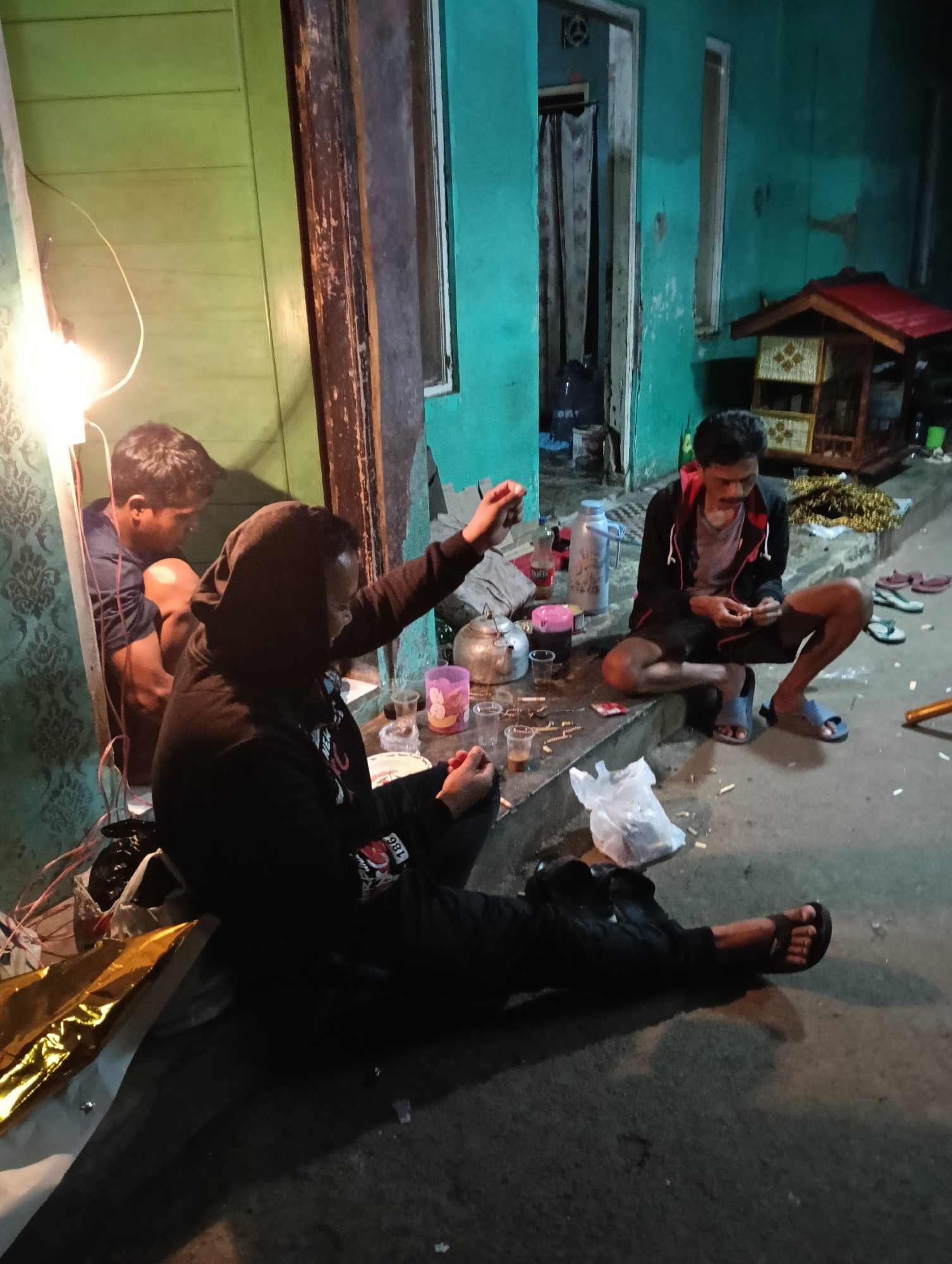 In Tasikmalaya, Indonesia, at night, three individuals are gathered on a paved outdoor area, likely a sidewalk or patio, adjacent to a building. The building has teal-colored walls and a green wooden door. A bright, bare lightbulb hangs from a wire, illuminating the immediate area.
In the foreground, a person wearing a black hooded sweatshirt and black pants is seated on the ground, facing away from the camera. Their left leg is extended, and their right leg is bent. They are holding an object in their right hand.
To the left of this person, a second individual, wearing a dark blue shirt, is crouched down, facing towards the right. They are partially obscured by the person in the hooded sweatshirt.
To the right, a third person is seated on a low concrete step. They are wearing a black jacket with red trim over a gray t-shirt and black shorts. They are looking down and appear to be handling small objects with their hands.
On the concrete step in front of the seated individuals, there are several items: a metal teapot, multiple clear plastic cups, some containing a dark liquid, a pink tumbler, and a bottle with a red label. A white plastic bag and various other debris, including what appear to be cigarette butts, are scattered on the ground. In the background, a small, ornate wooden structure, possibly a birdhouse or shrine, is visible. Several pairs of flip-flops are also present on the ground. The overall scene suggests a casual gathering.