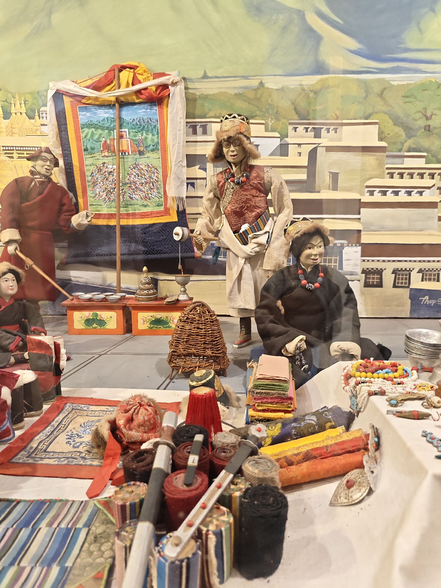 A diorama depicts a Tibetan scene. Three figures wearing traditional Tibetan clothing are in the foreground. The central figure wears a hat with fur trim and a vest over a light-colored robe. The figure to the left wears a red robe. The figure on the right wears a dark robe and sits cross-legged. A banner stands behind the figures, displaying a painting of a landscape with figures in traditional clothing. In front of the figures are various objects, including stacks of fabric, beads, and other items. The background of the diorama is a painting of a cityscape.