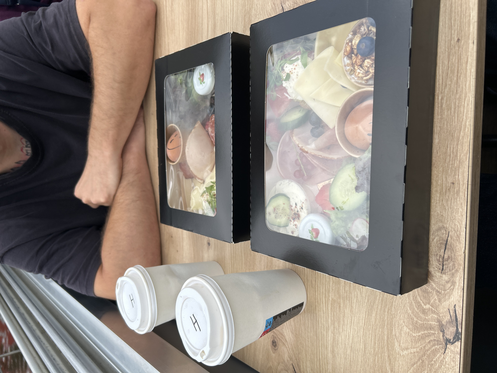 In an outdoor setting, possibly a cafe or restaurant with a wooden table, a person wearing a dark t-shirt is seated with their arms crossed. In front of them, two black takeout boxes with clear plastic lids are placed on the table, filled with various food items. The box in the foreground contains a selection of cheeses, cured meats, fruits like strawberries and blueberries, and what appears to be a creamy spread. The box behind it holds similar items, including sliced meats and small containers with garnishes. Two white paper coffee cups with lids, one with an "H" marking, are also on the table. The overall scene suggests a casual meal or snack outdoors.
