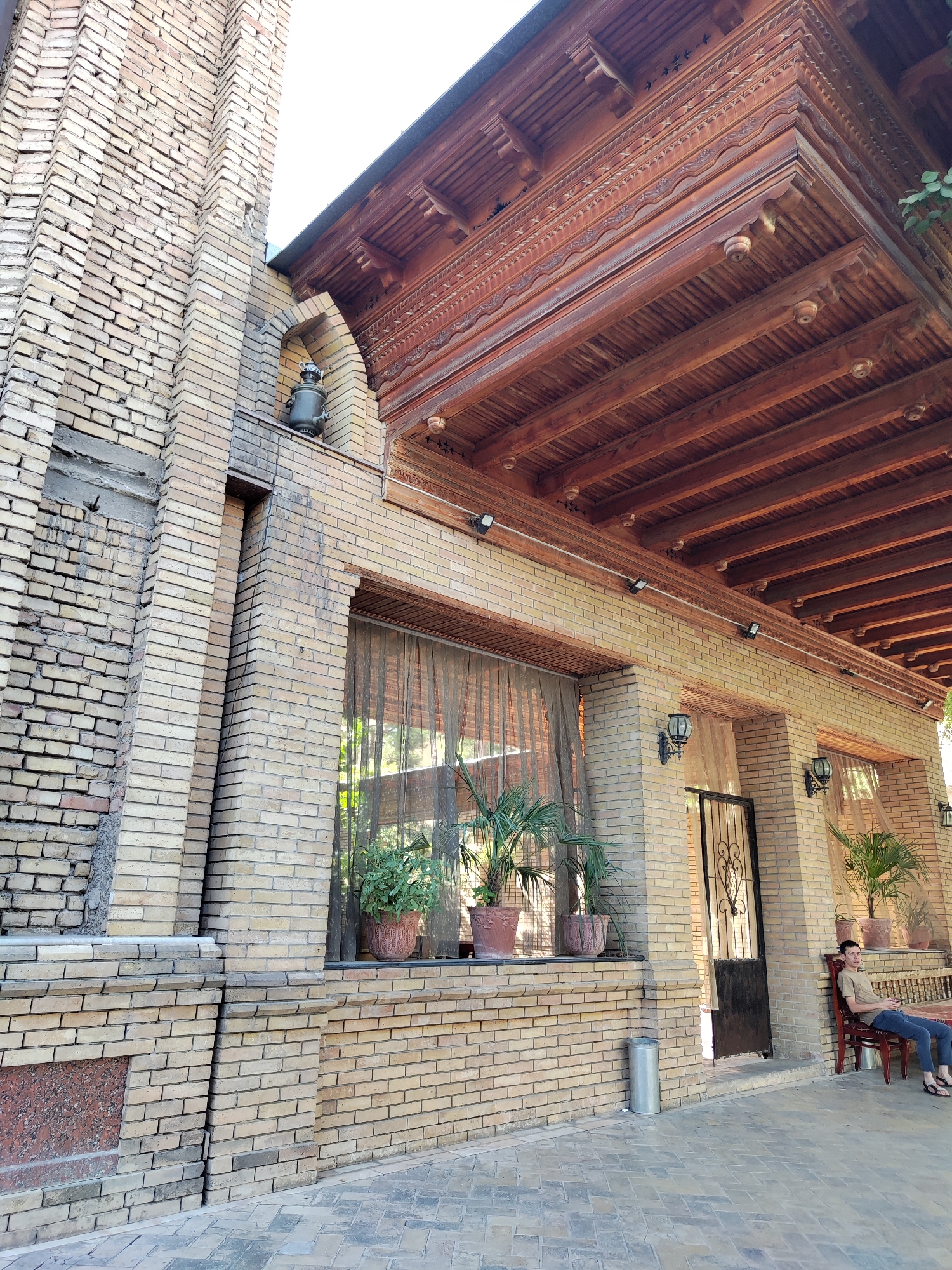 The image depicts the exterior of a building in Tashkent, Uzbekistan. The architecture features light-colored brickwork for the walls and a prominent wooden roof with intricate carvings. A tall, narrow brick column rises on the left side of the frame, adorned with a decorative niche containing a samovar. Large windows with sheer curtains are visible along the building's facade, with potted plants placed on a ledge beneath them. In the foreground, a person is seated on a wooden chair to the right, looking towards the left. The ground is paved with textured tiles. The overall scene suggests an outdoor seating area or entrance to a building with traditional architectural elements.
