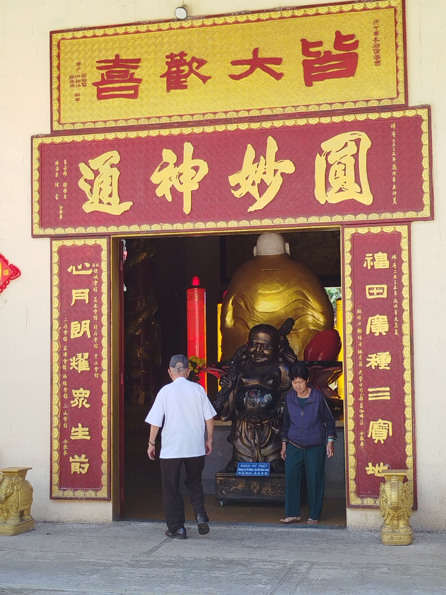 The image shows the entrance to a Buddhist temple in Da Lat, Vietnam. Two elderly individuals are present; an elderly man in a white shirt and dark pants is walking into the temple, while an elderly woman in a blue jacket and green pants is walking out. Inside the temple is a large golden Buddha statue with a smiling face. The entrance is framed by ornate, red and gold signs with Chinese characters. Above the entrance, two large signs are displayed, also with Chinese characters in gold on red and gold backgrounds. Flanking the doorway are two gold elephant statues serving as pedestals. The temple's exterior wall is painted off-white.