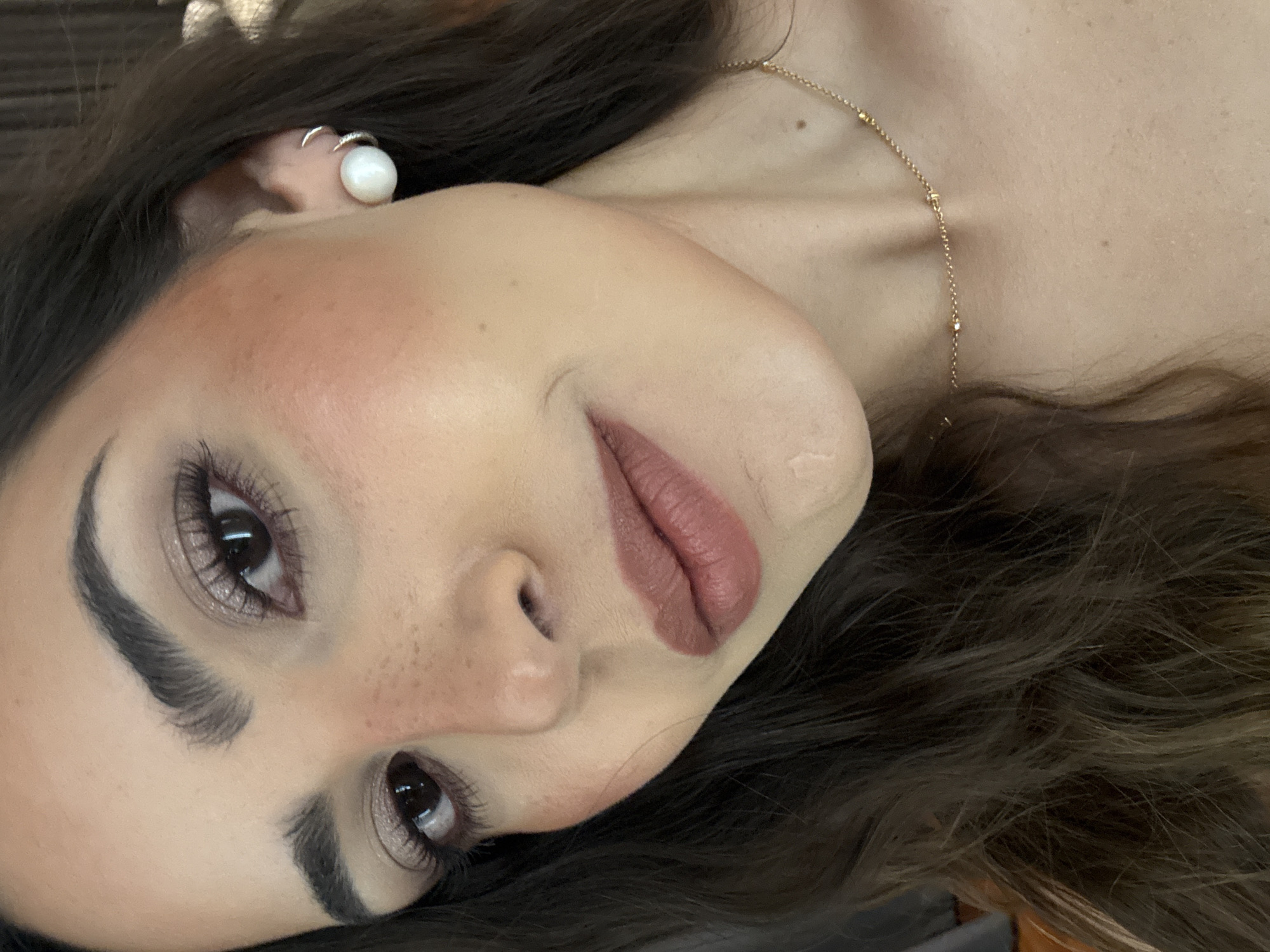 A close-up, angled headshot of a woman with fair skin. She has dark brown hair, full eyebrows, and brown eyes. Her makeup includes blush and lipstick. She is wearing a pearl earring in her left ear and a gold chain necklace. The background is indistinct. There is no information given in the prompt or image to suggest any relation to San Bernardino Tlaxcalancingo, Mexico.