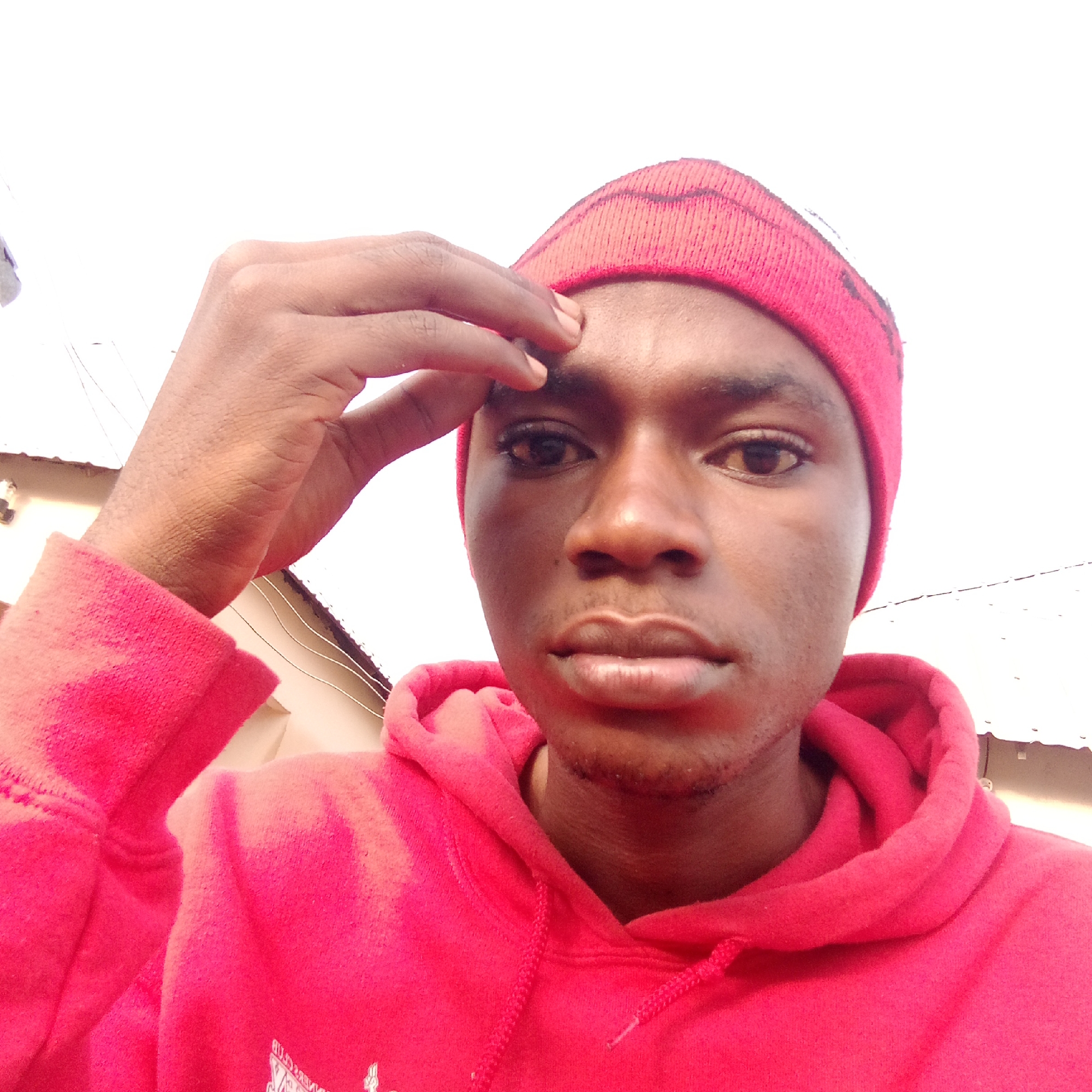 A close-up, outdoor selfie of a young Black man wearing a red hooded sweatshirt and a red beanie. He is holding his fingers to his forehead. Overhead electrical wires and the corner of a building are partially visible in the background. The location is suggested to be Mararaba, Nigeria.