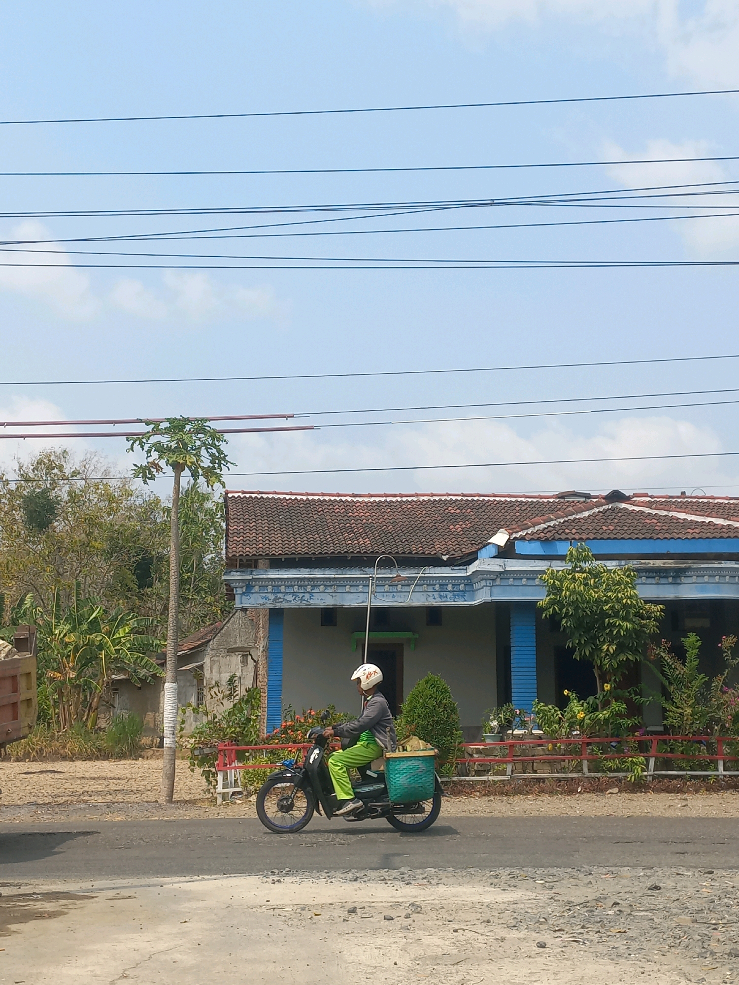 A person wearing a helmet and a grey jacket with green pants rides a black motorcycle on a paved road in Baturetno, Indonesia. The motorcycle has a green woven basket secured to the rear. In the background, a house with a red tile roof and blue trim is visible, with trees and foliage surrounding it. Overhead, power lines stretch across a light blue sky with scattered clouds. To the left of the motorcycle, a portion of a truck bed filled with sand is visible. The road surface is asphalt with scattered gravel.