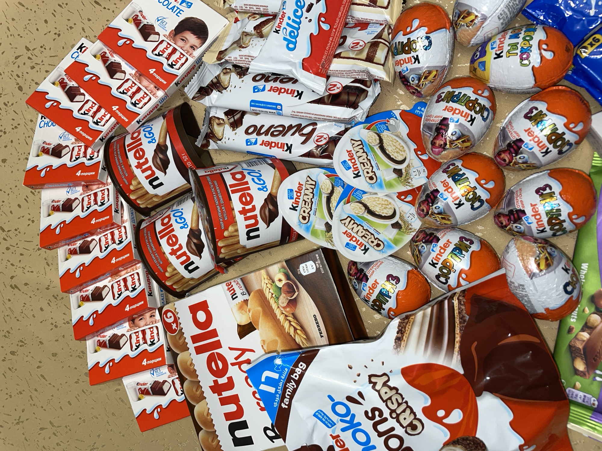 The image shows a variety of Kinder chocolate products scattered on a surface. The products include Kinder Chocolate bars, Kinder Bueno bars, Kinder Delice, Kinder Creamy, Kinder Joy eggs, Nutella & GO!, Nutella B-ready, and Kinder Crispy. The products are arranged haphazardly. Some packaging includes text in Russian or Arabic, indicating diverse markets. The location is not discernible from the image. There is no indication of Aratashen, Armenia.