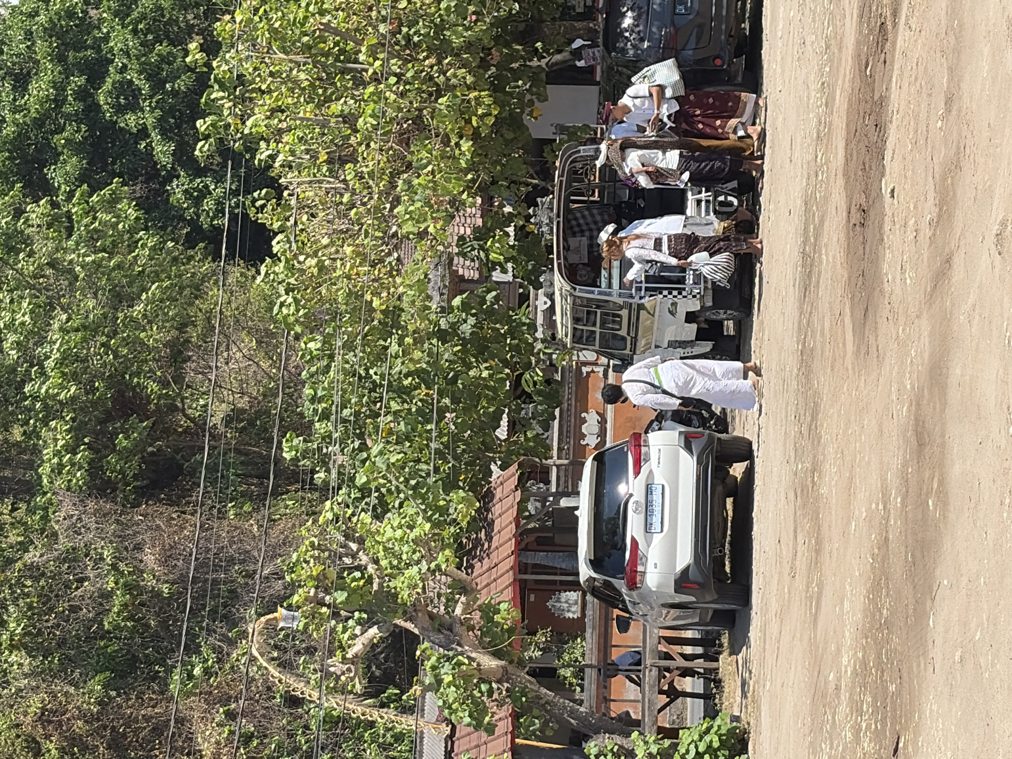 In Nusa Penida, Indonesia, several people dressed in traditional Balinese attire stand near a parked white SUV and a light-colored van. The individuals are observed in various poses, with some appearing to be engaged in conversation or preparing to depart. The white SUV is positioned on the left side of the image, its rear facing the viewer. The van, a more open-air vehicle, is situated to the right of the SUV. The ground is a dirt surface, indicative of an outdoor parking area. Behind the vehicles, a traditional Balinese building with a terracotta tile roof is visible, partially obscured by lush green trees. Power lines are strung across the scene, suggesting the presence of infrastructure. The overall environment suggests a warm, sunny day in a tropical location.
