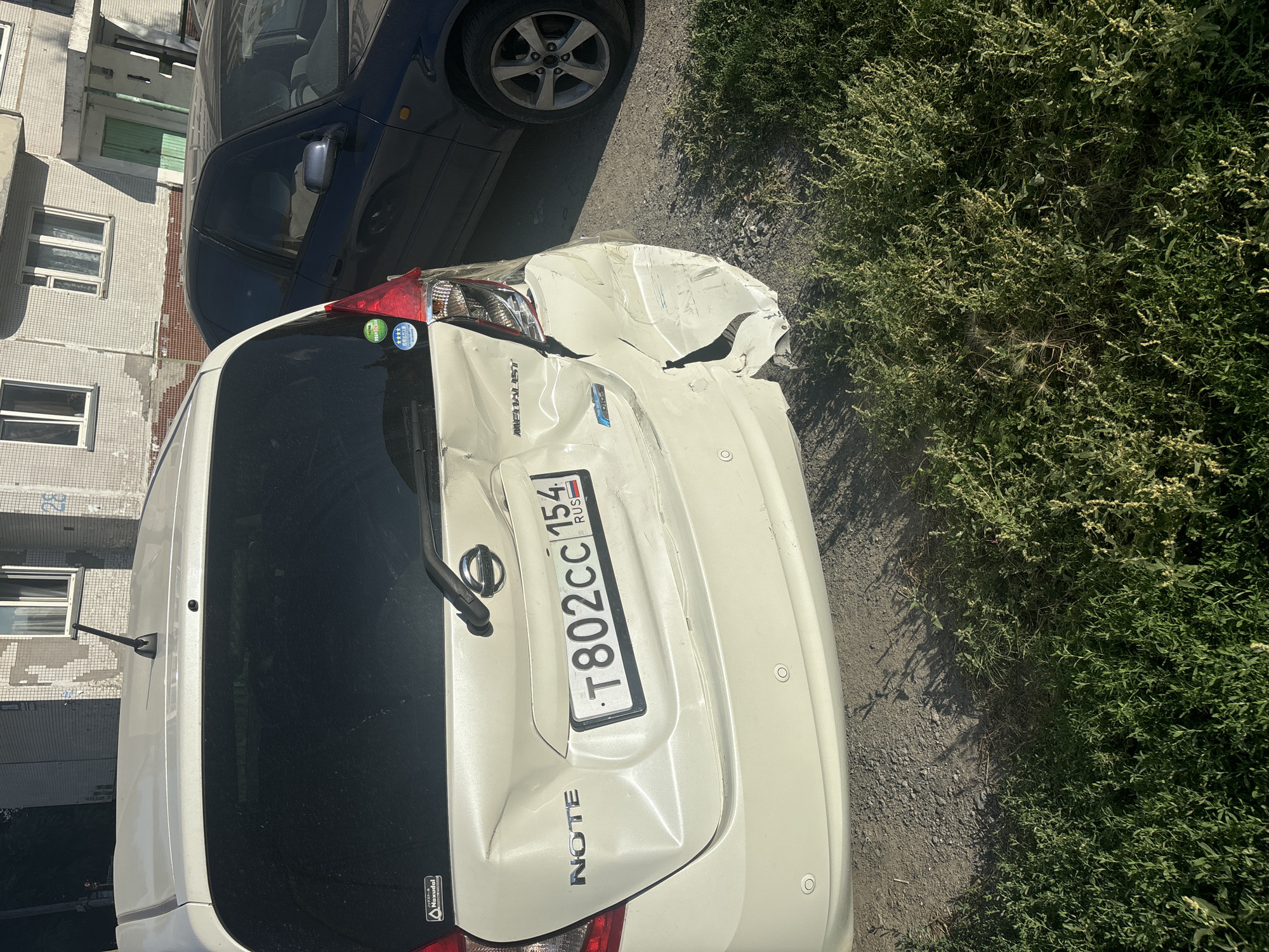 A white Nissan Note hatchback, license plate T802CC154 RUS, is parked on an asphalt surface next to a building in the city of Ob, Russia. The rear of the car is damaged on the right side, with significant crumpling and a large tear in the bumper and quarter panel. The car has a rear wiper and a sticker on the left side of the rear window. A dark-colored sedan is parked to the right of the Nissan Note. The building behind the cars appears to be residential, with a light-colored tiled facade and multiple windows. Green bushes are growing in the foreground.