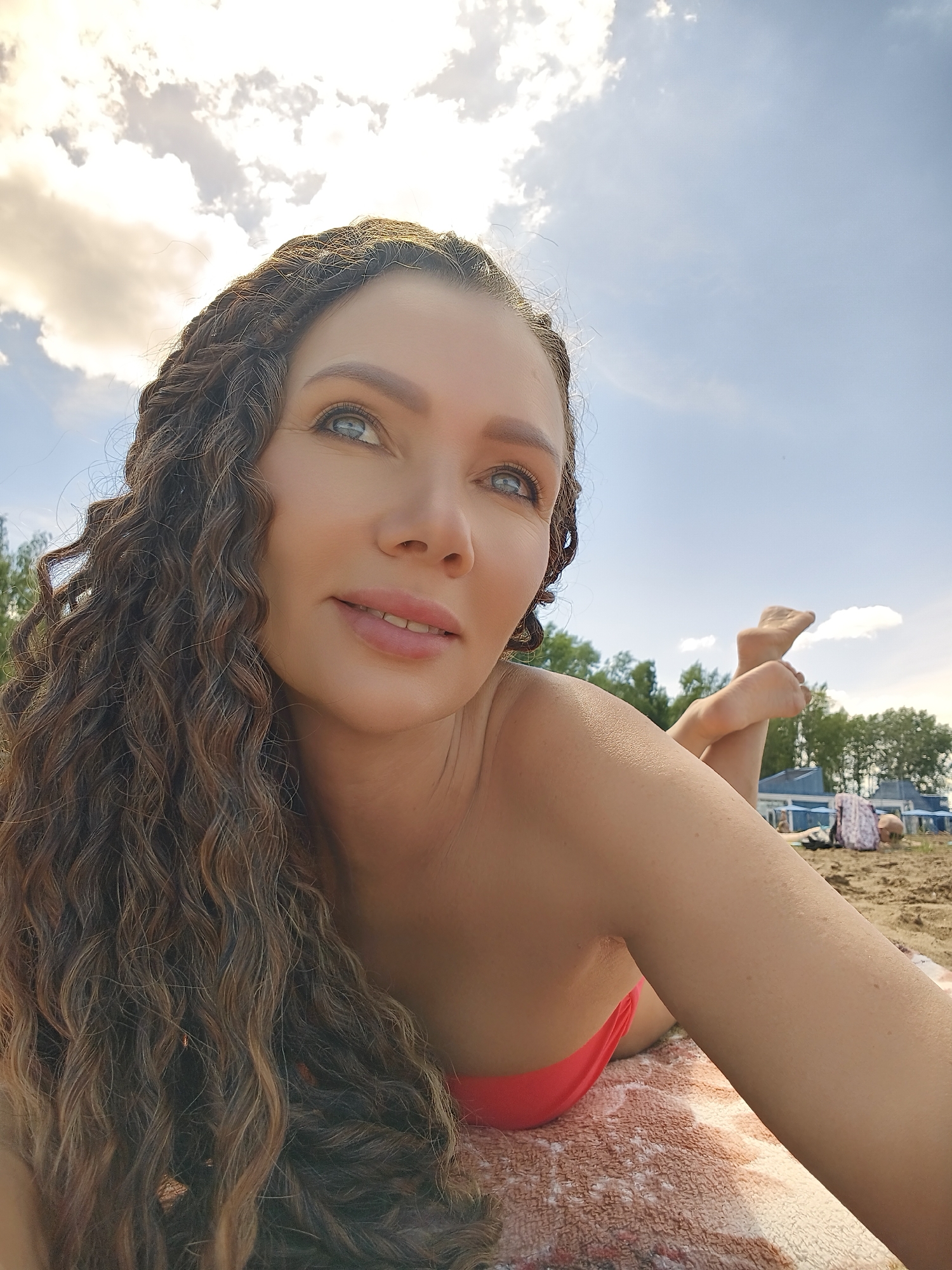 A woman with long, curly, braided brown hair lies on a sandy beach, looking up and to her right. She is wearing a pink bikini top. Her skin is tanned, and she has light blue eyes. She is lying on a patterned towel. Her feet are visible in the background, with one leg bent and resting on the sand. Other people are visible in the distance, along with some beach structures that appear to be cabanas or changing rooms. Trees are visible behind the beach structures, and the sky is blue with some white, fluffy clouds, suggesting it is daytime with pleasant weather. The lighting suggests it is either morning or late afternoon. There is no visible text in the image.