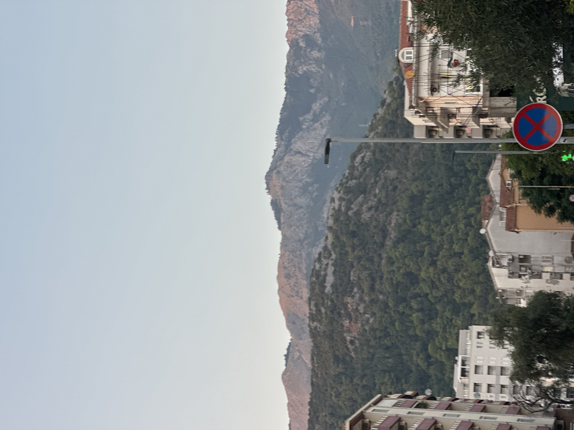 The image depicts a cityscape in Budva, Montenegro, dominated by layered mountains in the background. The sky is clear and pale blue, transitioning to a lighter hue near the horizon. In the foreground, several multi-story buildings are visible, characterized by balconies and apartment structures. A street light stands prominently in the mid-ground, and a round traffic sign with a red border, a blue background, and a red "X" is positioned on a pole. The vegetation consists of dense green trees covering the lower slopes of the mountains and scattered trees in the urban area. The lighting suggests it is either early morning or late afternoon, with a soft light illuminating the scene.