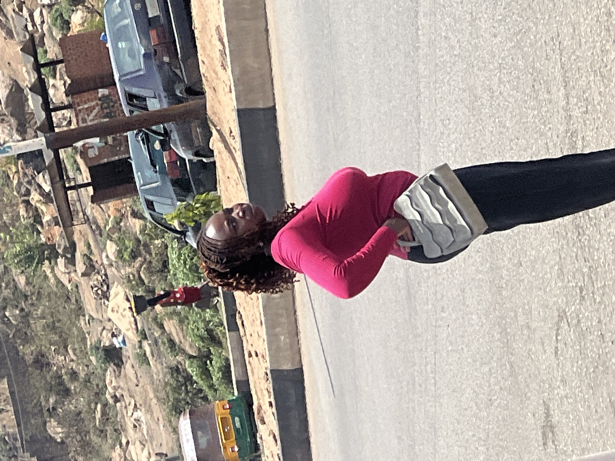 The image, captured in bright daylight, shows an outdoor scene, likely a street or road in Jos, Nigeria. The main subject is a woman with dark skin and braided hair, walking on the right side of an asphalt road. She wears a bright pink long-sleeved top, dark-colored pants, and carries a light-colored bag in her left hand. Her expression appears neutral as she looks slightly upwards.
To the woman's left, behind a curb, lies a rocky and hilly landscape with sparse green vegetation. Two older model cars, one blue/grey and one darker, are parked along the roadside. Behind the parked cars, a reddish-brown brick building is visible, along with a utility pole.
Further in the background, nestled within the rocky terrain, are several smaller figures, including one person in a red top. On a road in the distance, a yellow and green tricycle taxi (auto-rickshaw) is visible, a common sight in Nigeria. The weather is sunny and clear, casting distinct shadows. No clearly legible text is discernible on any of the vehicles or structures. The overall scene portrays a glimpse of daily life in a semi-urban or rural Nigerian setting.