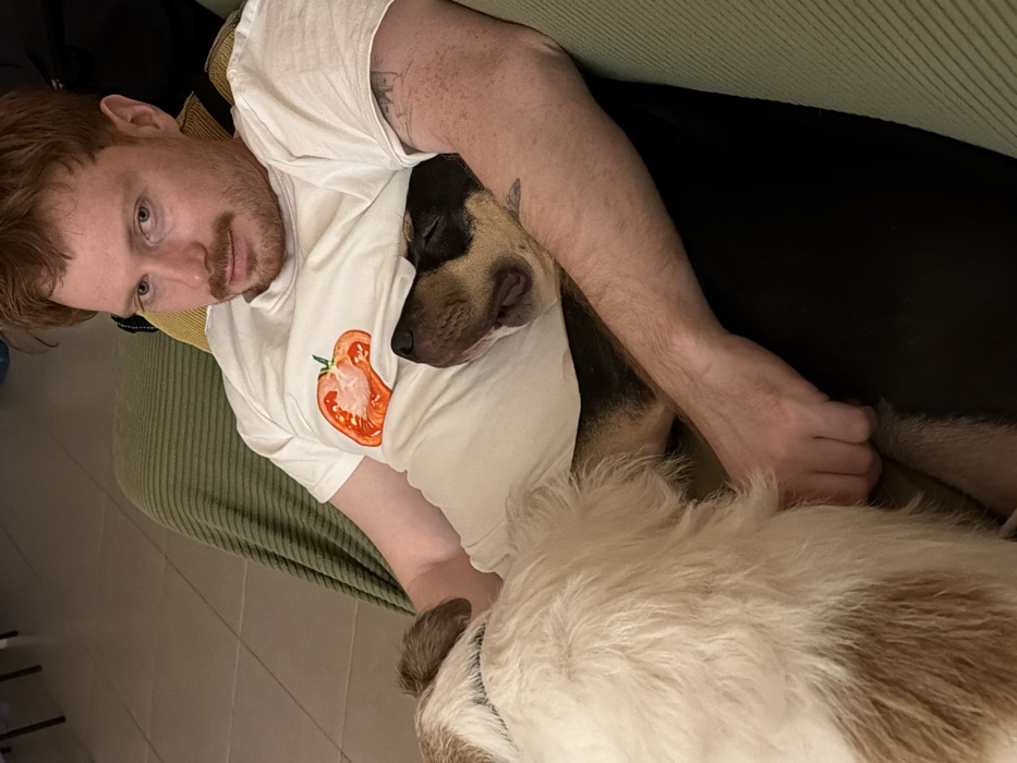 A man with reddish-brown hair and a light beard lies on a green corduroy couch, wearing a white t-shirt with a tomato graphic. He's cuddling with two dogs. One dog, with black and tan fur, nestles close to his chest. The other dog, with long white and brown fur, is positioned below. The man's arm rests gently on the dogs. The scene is lit naturally, creating soft shadows.