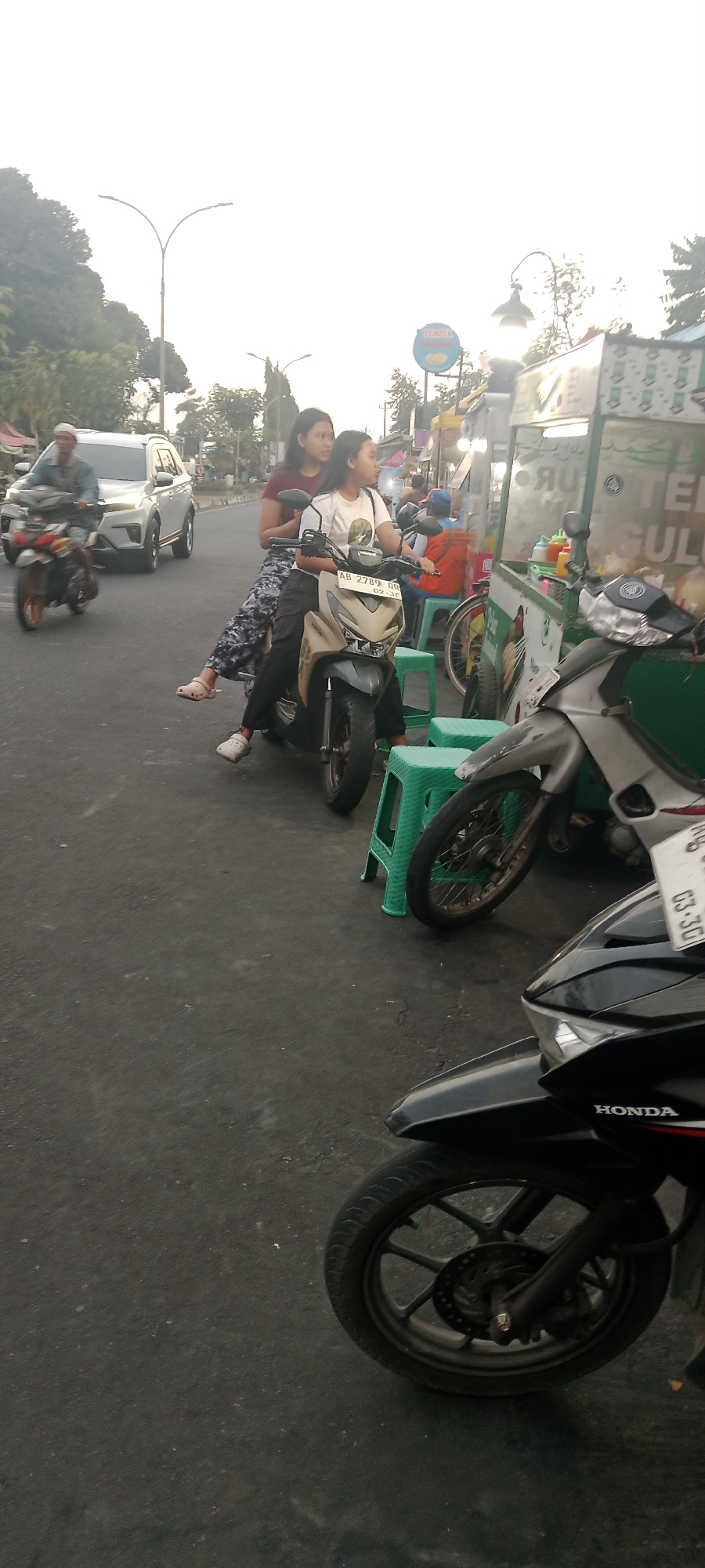 In the city of Ngemplak, Indonesia, a medium shot captures a street scene where two young women ride a scooter on a paved road. The woman in front, wearing a white t-shirt with a graphic print and black pants, operates the scooter. Behind her, another woman in a maroon t-shirt and patterned leggings sits as a passenger. Both are wearing casual footwear. The scooter itself is a beige and dark gray model with license plate "AB 2789 GR".
To the right of the scooter, several green plastic stools are visible, some of which are occupied by people. A food cart, adorned with green and white paneling and displaying various condiments, is positioned next to the stools. The cart appears to be selling food or beverages. In the background, a silver SUV is seen driving away on the left side of the road, and a man in a blue shirt and a white cap rides a motorcycle in the far left distance. The environment suggests a bustling commercial area with street vendors and moderate traffic. The sky above is bright and overcast, indicating daytime.