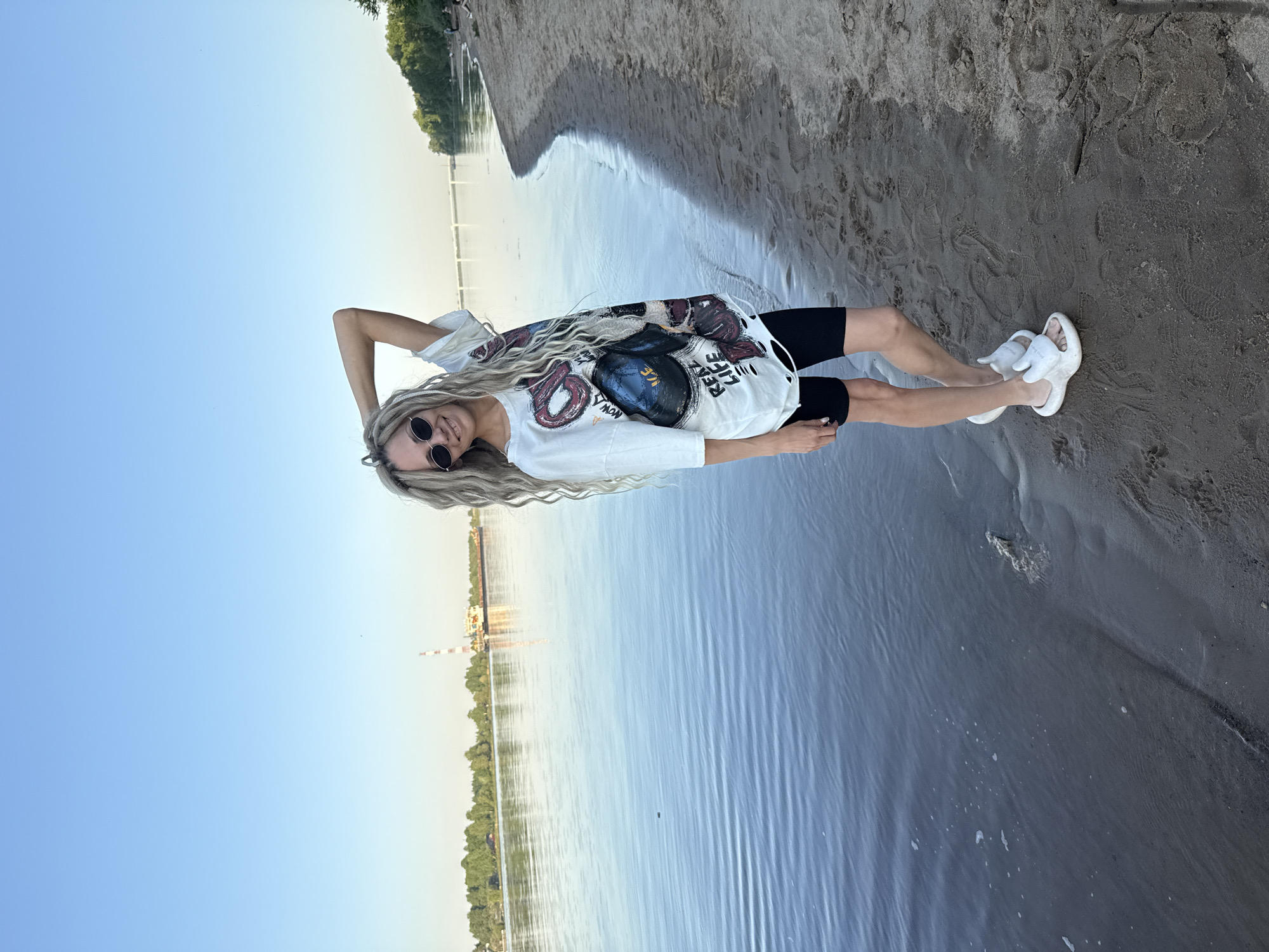 A woman with blonde dreadlocks is lying on a sandy beach with one arm raised above her head. She is wearing sunglasses, a white graphic tee, black shorts, and white sandals. The beach is bordered by water on one side and foliage on the other. In the distance, a pier, a shoreline with trees, and a tall industrial structure can be seen across the water. The location is Rusanovo, Russia.