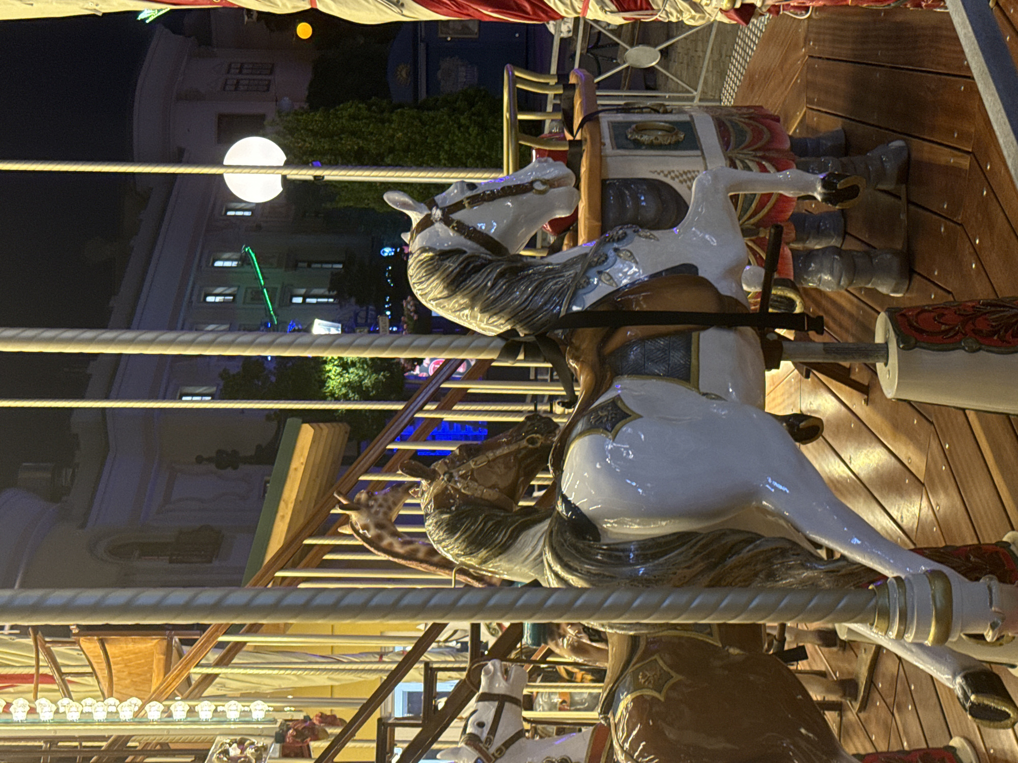 This is a night-time, medium-shot photograph of a carousel in Vienna, Austria. The carousel features several painted horses, one of which is white with a dark mane and tail, and another is brown and white. Behind the horses, a giraffe figure is visible. The carousel platform is made of polished wooden planks. In the background, a building with illuminated windows and a street lamp with a large spherical light are visible. The overall atmosphere is festive and nocturnal.