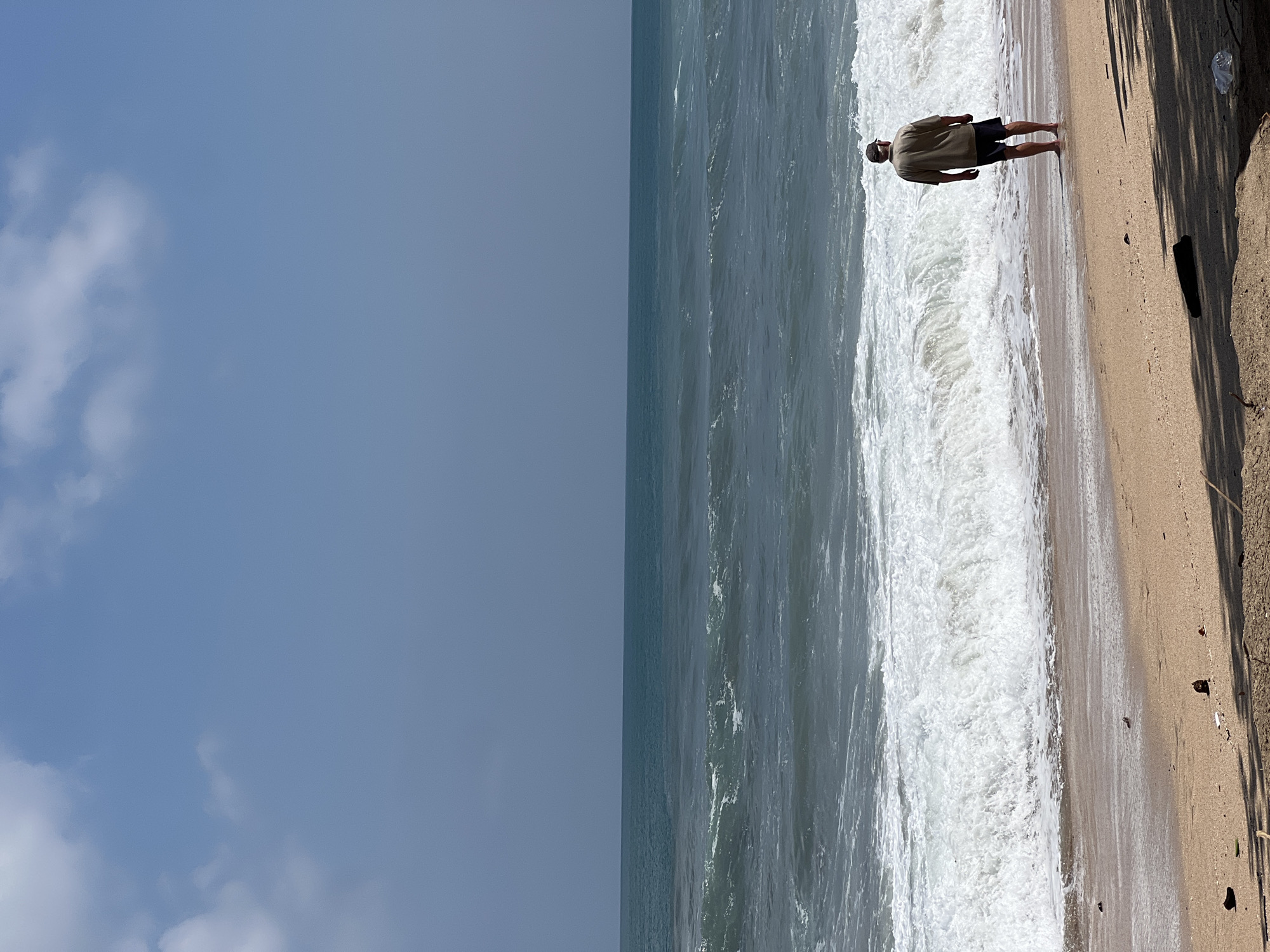 A lone person wearing a light brown shirt and dark shorts stands on a sandy beach in Kuta, Indonesia, facing the ocean. The person is facing away from the camera, with their back to the viewer, and their feet are submerged in the shallow water at the edge of the waves. The ocean is a deep blue-green with white foam from the crashing waves. The sky above is a clear blue with some scattered white clouds. The beach is sandy with some debris visible on the sand. The lighting suggests it is daytime, likely mid-morning or afternoon.