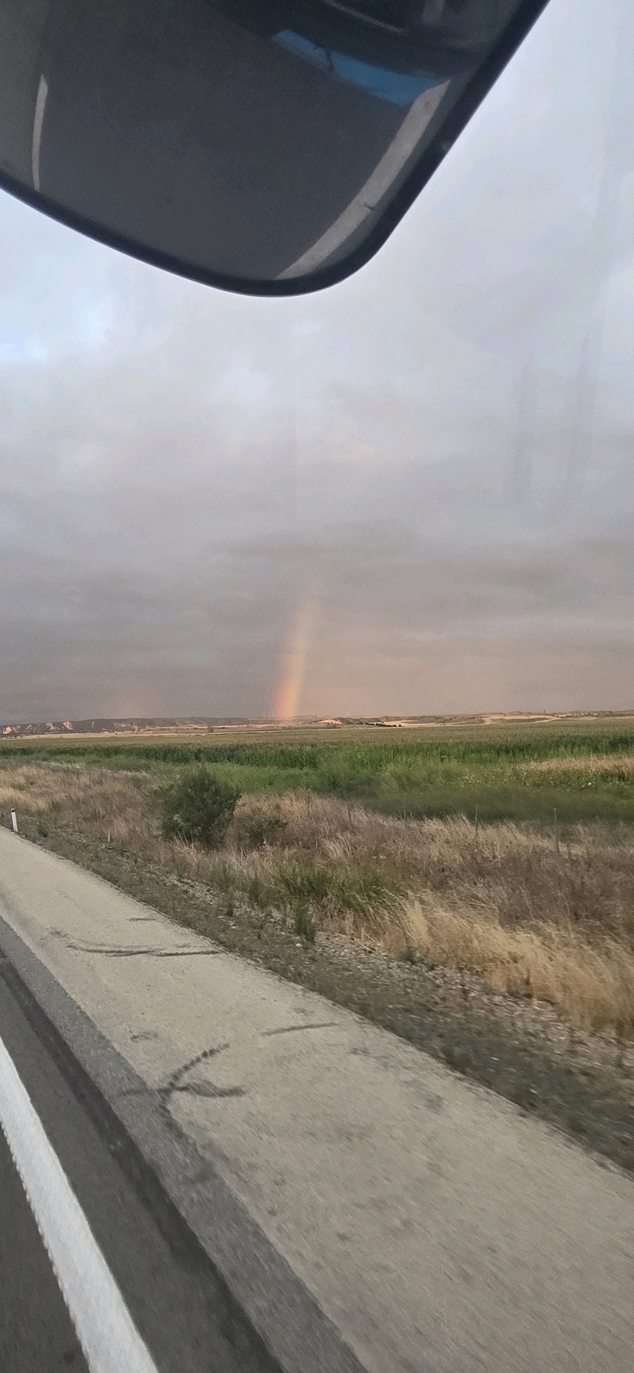 The image captures a view from a vehicle, looking out at a landscape under a cloudy sky. A faint rainbow arcs across the horizon in the distance. In the foreground, a paved road with white lane markings runs diagonally from the bottom left corner towards the center. The road appears to be slightly wet or stained in places. To the right of the road, a field of dry, golden-brown grass and some green vegetation stretches towards the horizon. Further back, a line of trees and agricultural fields are visible. The top portion of the image is dominated by the interior of the vehicle, with a dark gray panel and a sliver of blue visible, suggesting it's the car's interior or a reflection. The overall scene depicts a rural roadside view with a natural phenomenon, a rainbow, appearing after or during precipitation. The location is identified as the city of Marcilla, Spain.