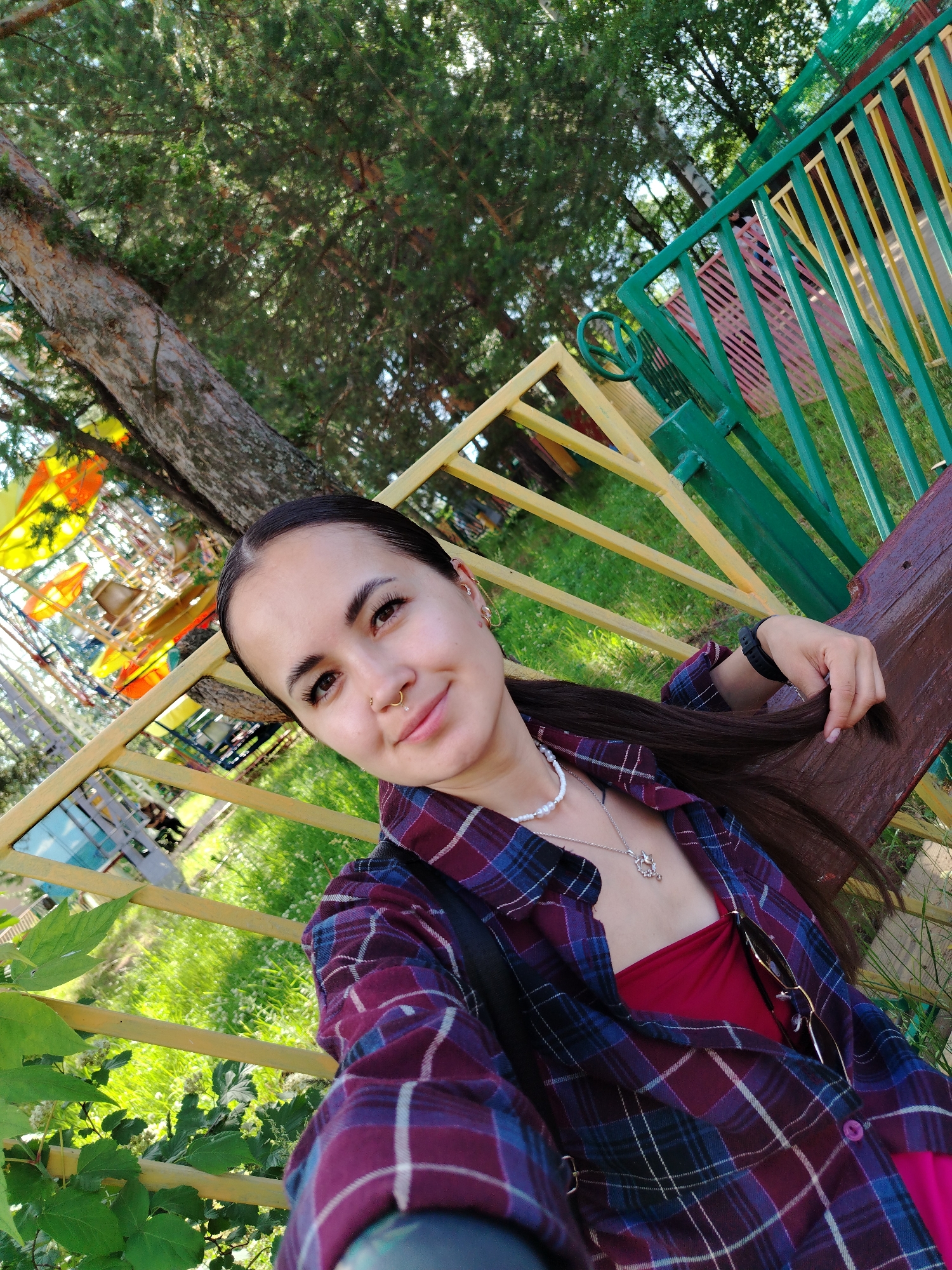 A young woman with dark hair pulled back and wearing a purple and blue plaid shirt over a maroon top, is taking a selfie outdoors. She has a nose ring and several earrings. She is sitting on a brown wooden bench in front of a yellow and green metal fence. In the background, trees, grass, and playground equipment are visible. The scene appears to be in an outdoor recreational area, possibly a park or playground in Chaikovsky, Russia.