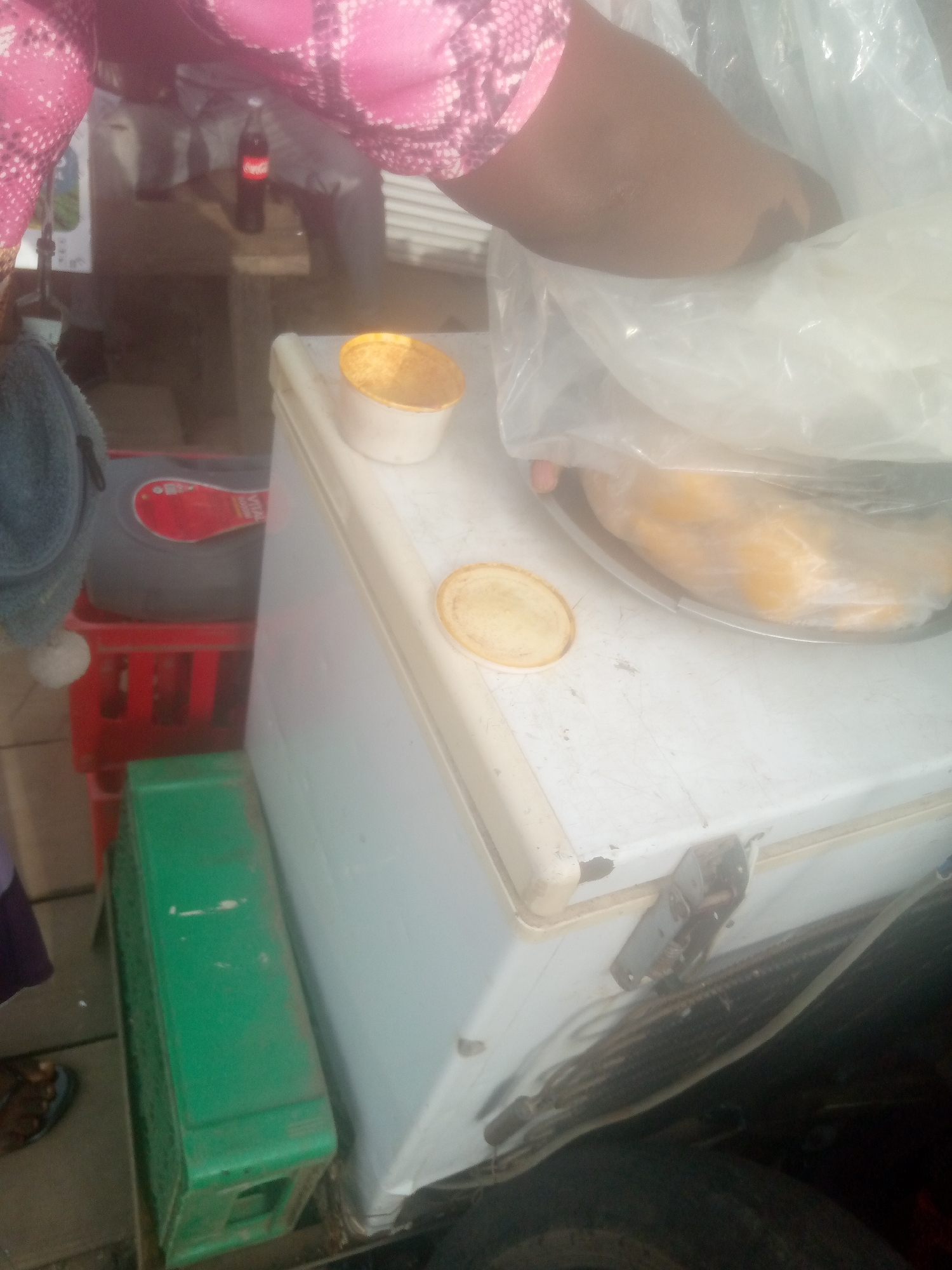 In a street scene in Jimeta, Nigeria, a person wearing a pink and white patterned garment and a brown arm is visible from the chest up, leaning over a white freezer. On top of the freezer, a small white cup with a golden lid is placed next to a clear plastic bag containing yellow items, possibly food. A red plastic basket filled with a grey object with a red label is to the left of the freezer, partially obscured by the person's arm and clothing. Below the red basket, a green plastic container is visible. The background is blurred, showing a table and a bottle of Coca-Cola. The overall scene suggests a commercial or informal market setting.