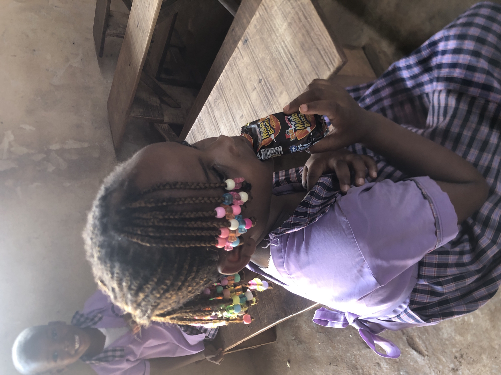 A young person, likely a girl, is shown in a close-up, angled shot, eating from a packet of biscuits. Her braided dark hair is adorned with numerous colorful beads in shades of pink, blue, white, and yellow. She wears a light purple short-sleeved top and a blue and white plaid skirt. She holds a black and orange packet of "Mallo Biscuits" or "MALTO Biscuits," with "PEANUT FLAVOUR" visible, along with a barcode and a Facebook logo.
She is positioned at a wooden table or desk. The floor is a light-colored, possibly concrete or dirt, surface. In the lower-left background, another young child, also in a purple top, is partially visible, smiling towards the camera. The scene is indoors, with ambient lighting suggesting daytime. The overall impression is a casual moment, possibly in a school or home environment in Ukwu-Abwa, Nigeria.