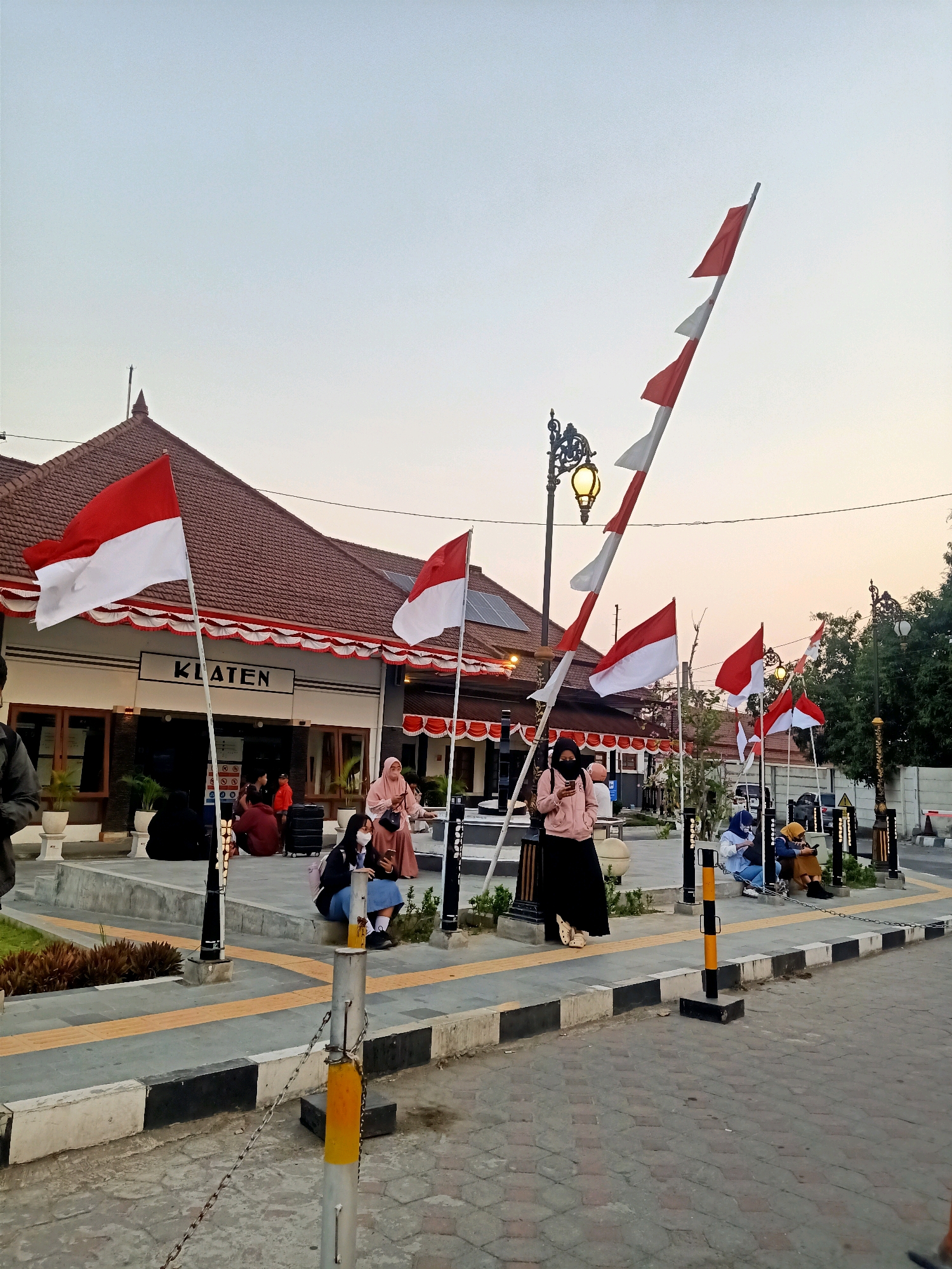 The image depicts the exterior of the Klaten train station in Klaten, Indonesia, during what appears to be late afternoon or early evening. Red and white Indonesian flags are prominently displayed, some flying from poles and others mounted on the building's facade. The building itself features a traditional tiled roof and a sign that reads "KLATEN" above the entrance. Several people are visible, some seated on a raised platform in front of the station, while others are standing. A few individuals are holding mobile phones. Decorative street lamps with a vintage design are also present, along with paved walkways and a brick-paved road. The sky is a pale blue, suggesting the sun is low on the horizon.