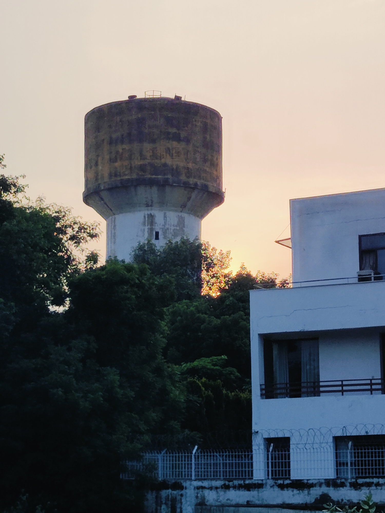 The image captures a water tower and a modern building in Gurgaon, India, against a soft, hazy sky suggesting dusk or dawn. The water tower, a cylindrical structure with a wider, weathered top section and a tapering base, stands prominently on the left. The top part of the tower exhibits visible discoloration and texture, suggesting age. A small rectangular opening is visible on the white base of the tower.
To the right of the water tower, a modern, multi-story building with white exterior walls and dark-framed windows is partially visible. The building has balconies with railings. A fence, possibly made of metal bars and barbed wire, runs along the foreground in front of the building. Lush green trees and foliage surround the water tower and the base of the building, creating a contrast between the natural elements and the urban structures. The soft light in the sky hints at the time of day. There are no people or discernible actions depicted in the image. The scene appears to be a quiet, perhaps residential or commercial, area in Gurgaon.