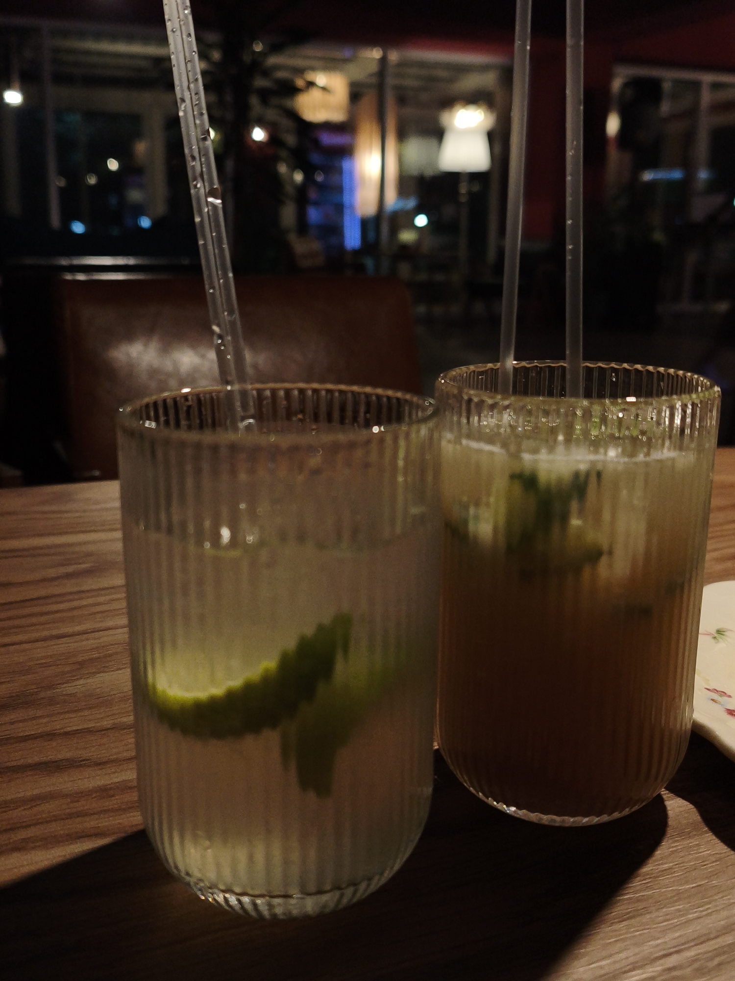 Two tall, ribbed glasses containing beverages with ice and lime wedges, each with a clear plastic straw, are positioned on a wooden table. The glass on the left contains a lighter-colored liquid with a slice of lime. The glass on the right holds a darker, amber-colored liquid with pieces of lime or mint visible within. The background is dimly lit and appears to be an indoor setting, possibly a restaurant or bar, with reflections of lights and furniture visible through a window or glass partition. There are no people visible in the image. The image is focused on the drinks.