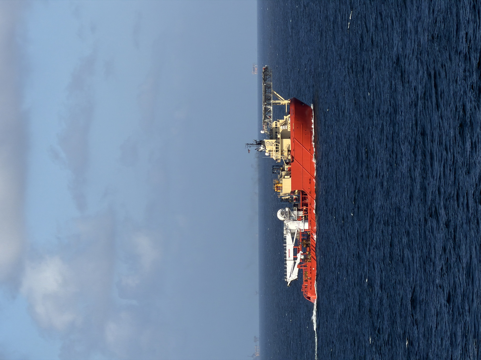 A large, red and white offshore support vessel is depicted underway on a blue ocean under a partly cloudy, light blue sky. The vessel is captured from an elevated perspective, showcasing its full length as it moves from left to right, leaving a distinct white wake behind its stern.
The ship features a complex superstructure, predominantly white, with various masts, antennas, and specialized equipment. A prominent white crane, extending over the deck, has the words "MAERSK" and "SUBSEA" visible on its side, identifying the vessel's operator and function. Further towards the stern, another structure bears the text "MAERSK SUPPLY". The hull of the ship is a vibrant red.
In the far distance, on the horizon line between the sea and sky, a smaller, indistinct vessel or offshore installation can be faintly seen. The sea surface is a deep blue with visible texture from small waves, suggesting moderate conditions. The sky is bright, indicating daytime, with scattered white and light gray clouds. The overall scene portrays an active industrial vessel engaged in maritime operations in open waters.